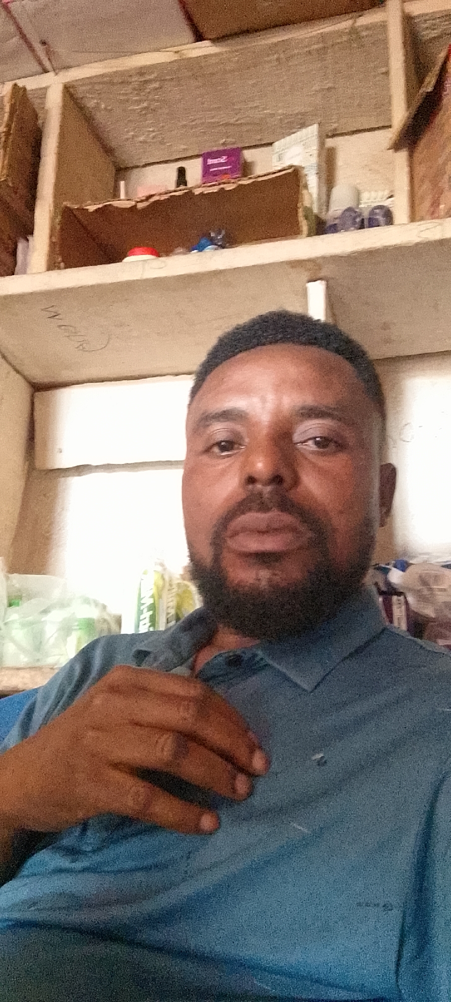 A medium shot captures a Black man with dark skin, a short beard, and short black hair, looking directly at the camera. He is wearing a blue collared shirt, and his right hand is resting on his chest. Behind him, there are shelves filled with various items, including boxes, plastic bags, and what appears to be a bottle. The background suggests an indoor environment, possibly a storeroom or workshop, with a rough, unfinished ceiling above the shelves. There is no discernible information about the location in Durumi, Nigeria.