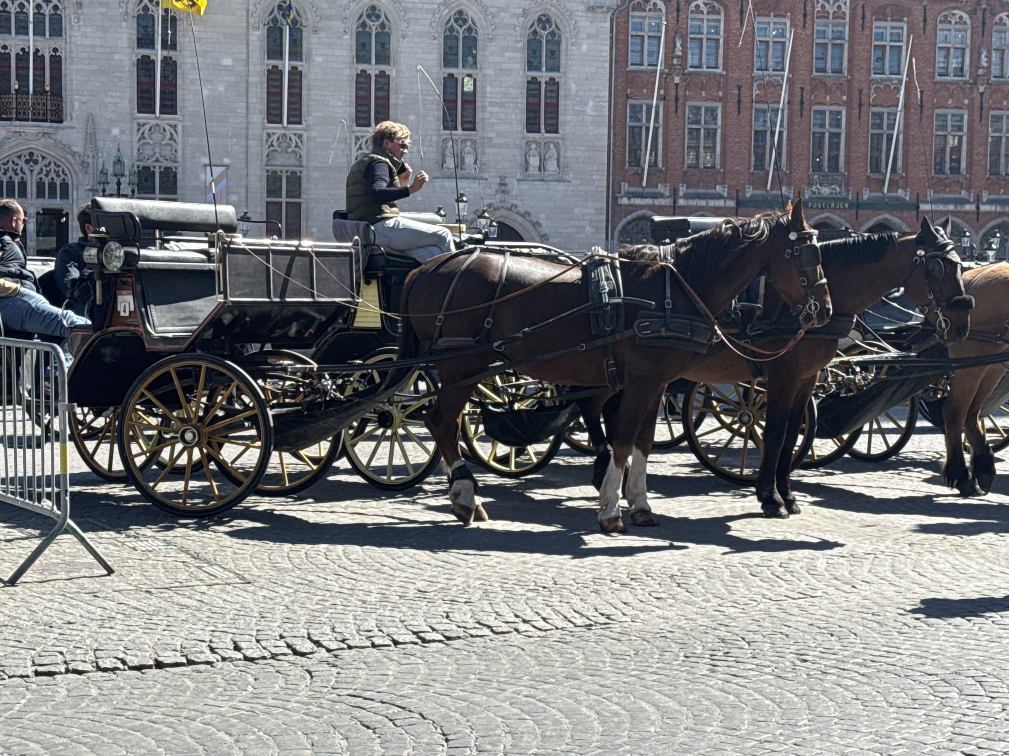The media file presents a sunlit scene in Bruges, Belgium, featuring a horse-drawn carriage. The primary subjects are two brown horses, harnessed to a traditional black carriage with gold-colored spoked wheels. A male driver, wearing a dark vest, light pants, and sunglasses, is seated on the carriage, bringing a small item to his mouth. A passenger in a dark jacket and blue jeans is visible on the left side of the carriage, resting his legs on a metal barrier.
The activity appears to be a pause during a horse-drawn carriage tour. The horses are standing still on a cobblestone street, suggesting they are waiting or taking a break.
Notable details include the bright, clear weather with strong shadows, indicating midday or early afternoon. In the background, there's an ornate Gothic-style stone building with arched windows and decorative sculptures, partially obscured by a yellow flag on a pole and a smaller, striped flag. To its right stands a red brick building featuring multiple windows and a visible sign that reads "POSTERIJEN," which translates to "Post Office" in Dutch. The street is paved with distinctive cobblestones, a common feature in historic European cities. A metal crowd control barrier is visible in the foreground on the left. The overall scene conveys a relaxed, tourist-oriented atmosphere.