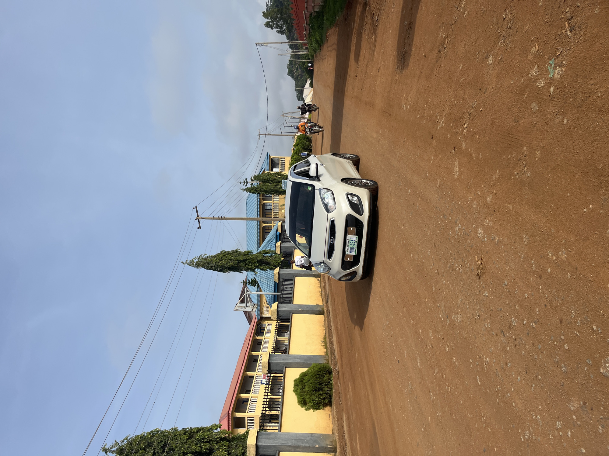 The image captures a street scene in Bukuru, Nigeria, on an overcast day. A white Kia Picanto car, with license plate MBD 494E, is the primary subject in the foreground, positioned on a dirt road. The road surface is reddish-brown and appears somewhat uneven.
To the left of the car, a yellow building with a red-tiled roof stands behind a black metal fence and a low yellow wall. Tall, green trees are planted along the fence line. To the right of the yellow building, a blue-roofed structure is visible, and further in the distance, a basketball hoop is mounted on a pole.
Several people are present in the scene. Two individuals are riding motorcycles in the middle ground, heading away from the viewer. One rider wears an orange shirt, and the other is in darker clothing. In the background, on the right side of the road, a few more figures are walking. Power lines are strung across the sky, supported by utility poles. The sky itself is a pale blue with scattered clouds. The overall environment suggests a semi-urban or developing area.