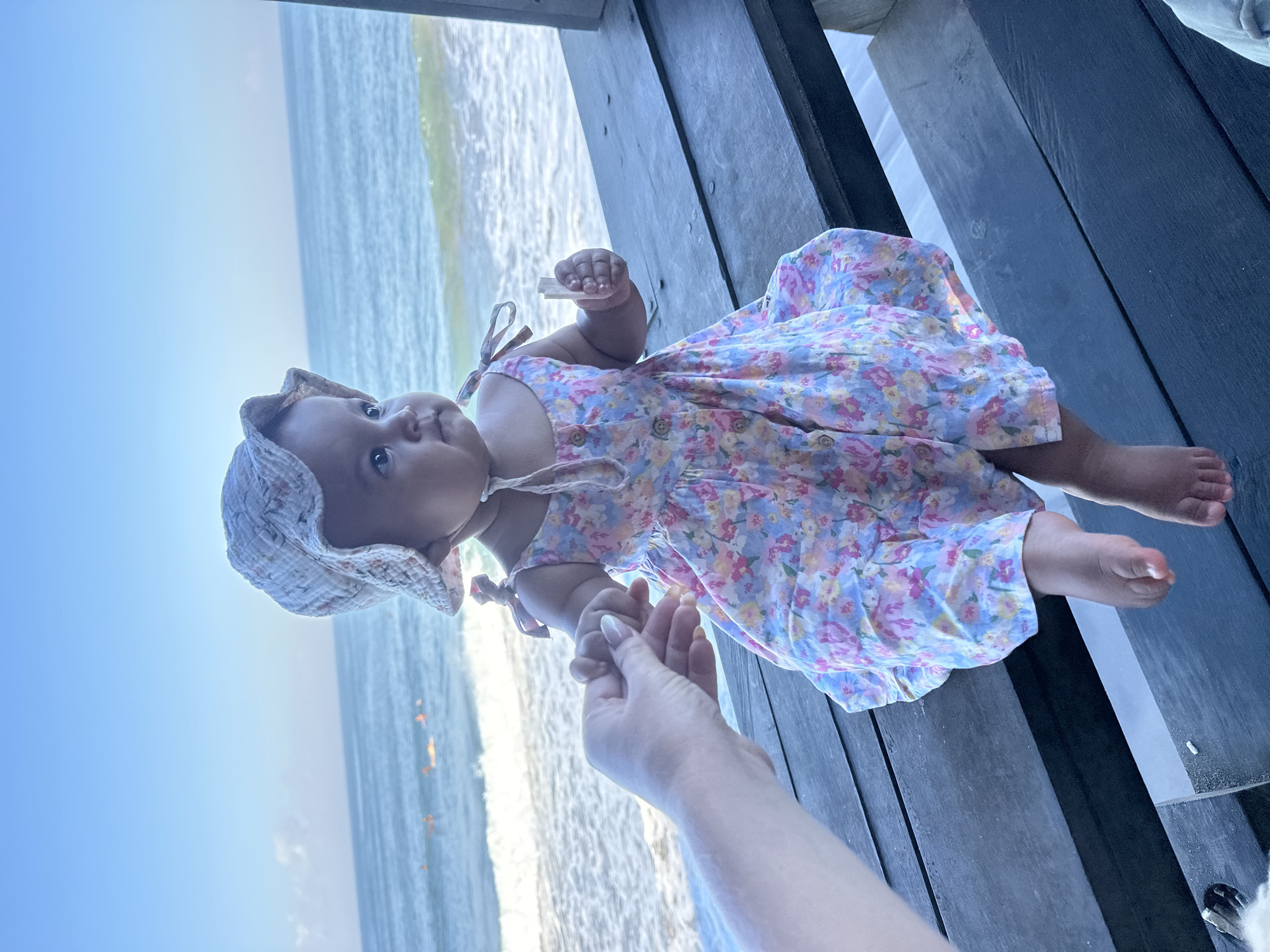 A baby wearing a floral print dress and a sun hat sits on a wooden bench facing the ocean in North Kuta, Indonesia. The baby's hand is held by an adult's hand, and the baby is holding a white object in their other hand. The ocean is visible in the background with waves and several people in the water.