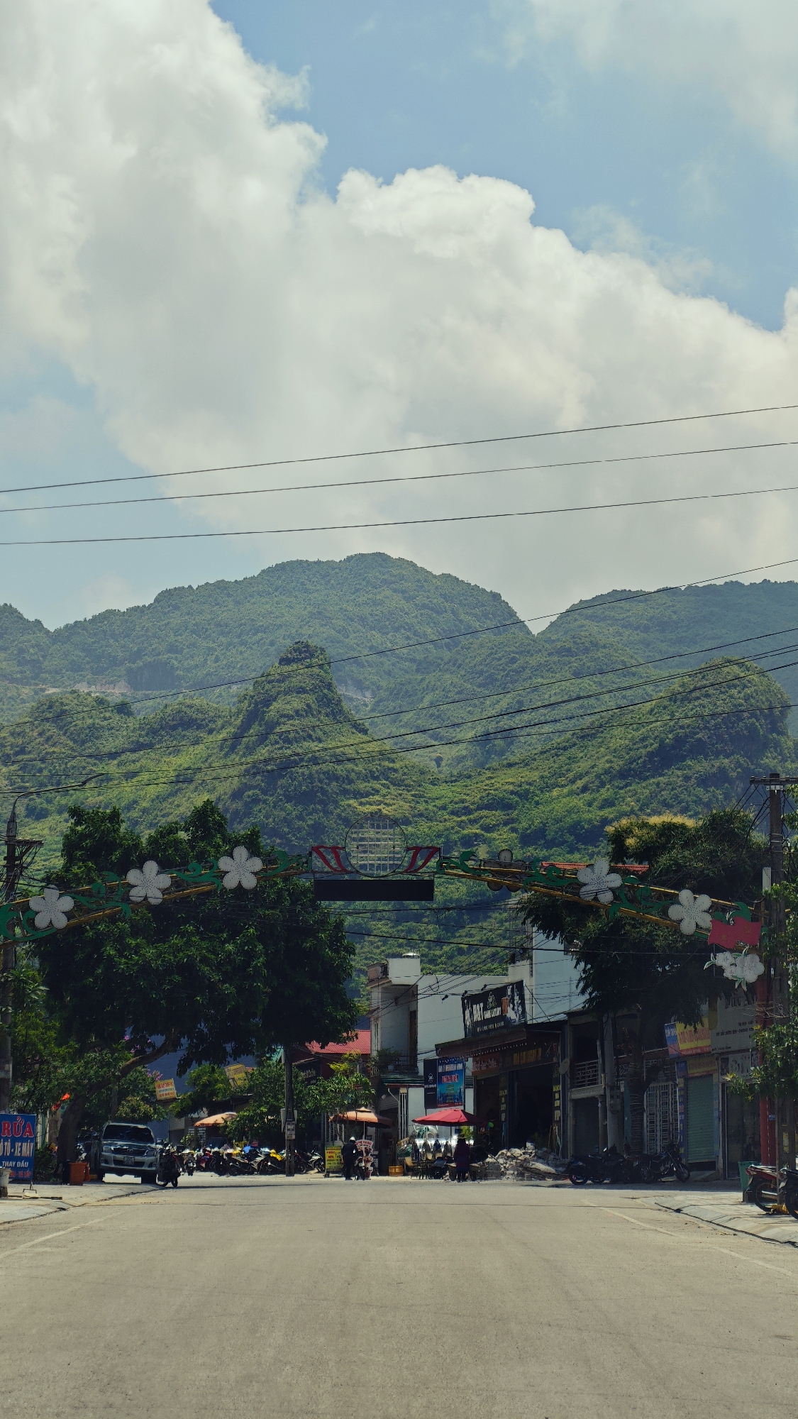 The image shows a street in Mèo Vạc Commune, Vietnam. The street is paved and leads towards green, mountainous terrain under a partly cloudy sky. Buildings line both sides of the street, with some having businesses operating from them, indicated by signs and awnings. Motorbikes and at least one car are parked on the sides. A decorative archway spans across the street, adorned with lights and flowers. Power lines are visible above the archway. There are a few people visible along the street.