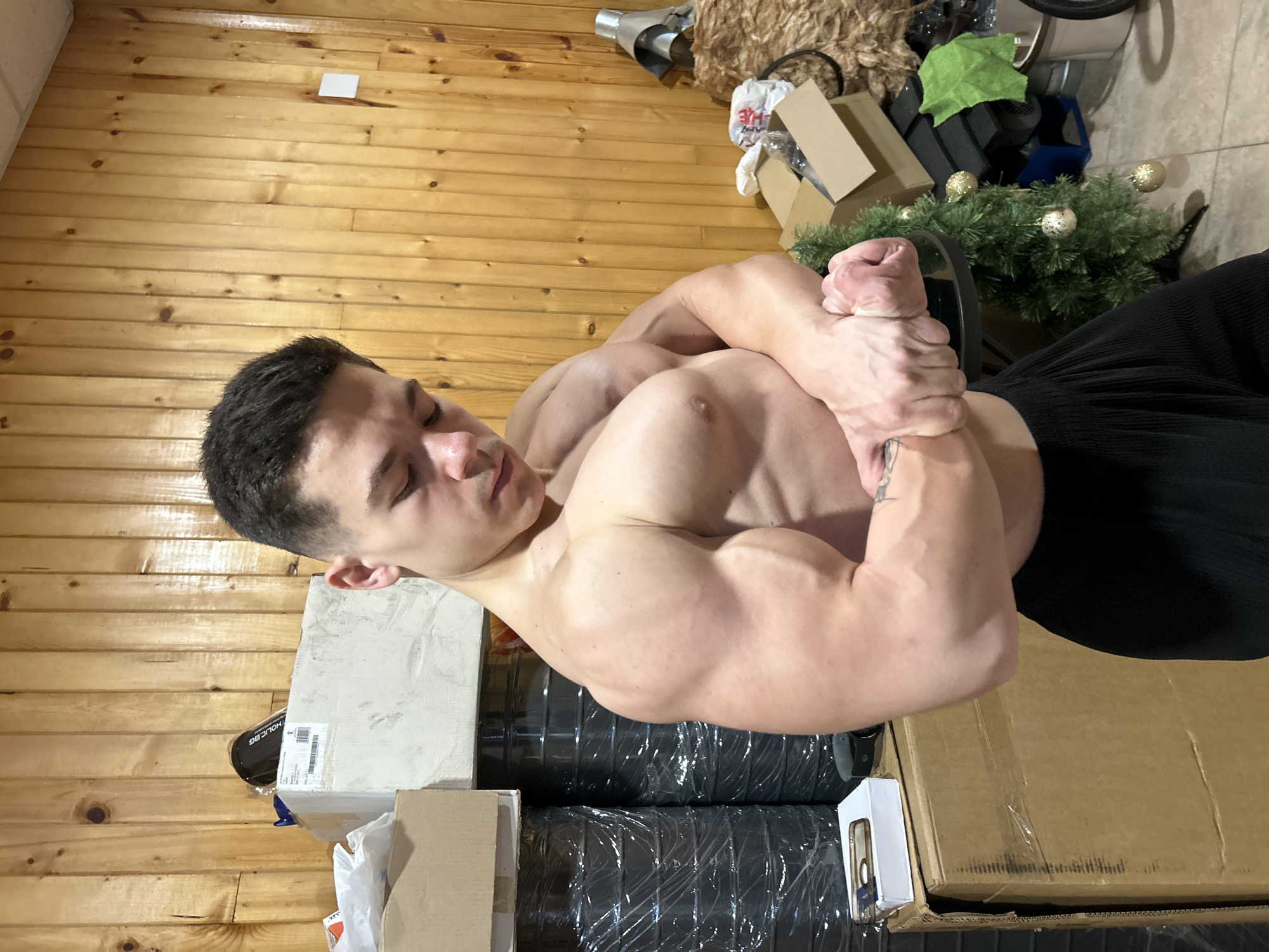 A muscular man is positioned in front of a wood-paneled wall. He is shirtless and wearing dark pants. His arms are flexed and clasped together. The floor appears to be tiled. There are boxes and other items surrounding the man, including a small decorated Christmas tree. The location is not discernable based on the image, so it is not possible to determine if it is in Troyan, Bulgaria.