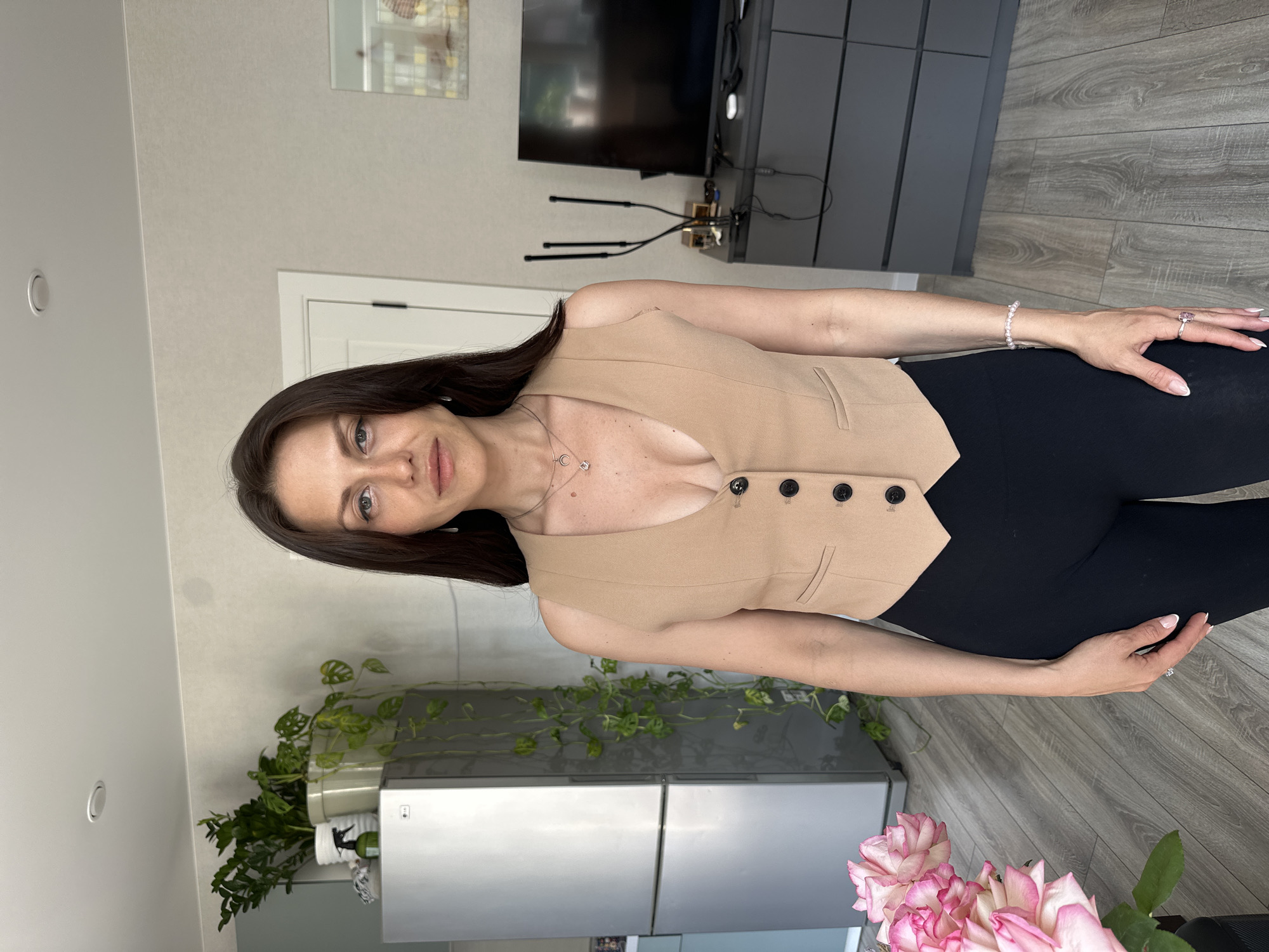 A full-body, waist-up shot captures a woman standing in an indoor setting that appears to be a residential apartment in Murino, Russia. The woman has long, dark brown hair and is facing the camera directly. She is wearing a tan, sleeveless vest with a deep V-neck over a black, high-waisted bottom. The vest features three black buttons down the front and a decorative pocket flap on the left side. Around her neck, she wears a delicate silver necklace with a small pendant. Her left arm is relaxed at her side, adorned with a thin, beaded bracelet and a ring on her ring finger.
The background reveals a moderately lit room. To the left, a silver LG refrigerator stands against a light-colored wall, with several potted plants of varying sizes situated on top of it. One plant, with long, trailing vines, hangs down beside the refrigerator. To the right, a dark gray dresser or cabinet is visible, topped with a black television. A decorative metallic object with multiple thin arms stands on the dresser, next to the TV. The flooring is a light wood laminate. In the foreground, to the left of the woman, a large bouquet of pink and white roses is partially visible. The overall environment suggests a domestic, possibly living room or kitchen area.
