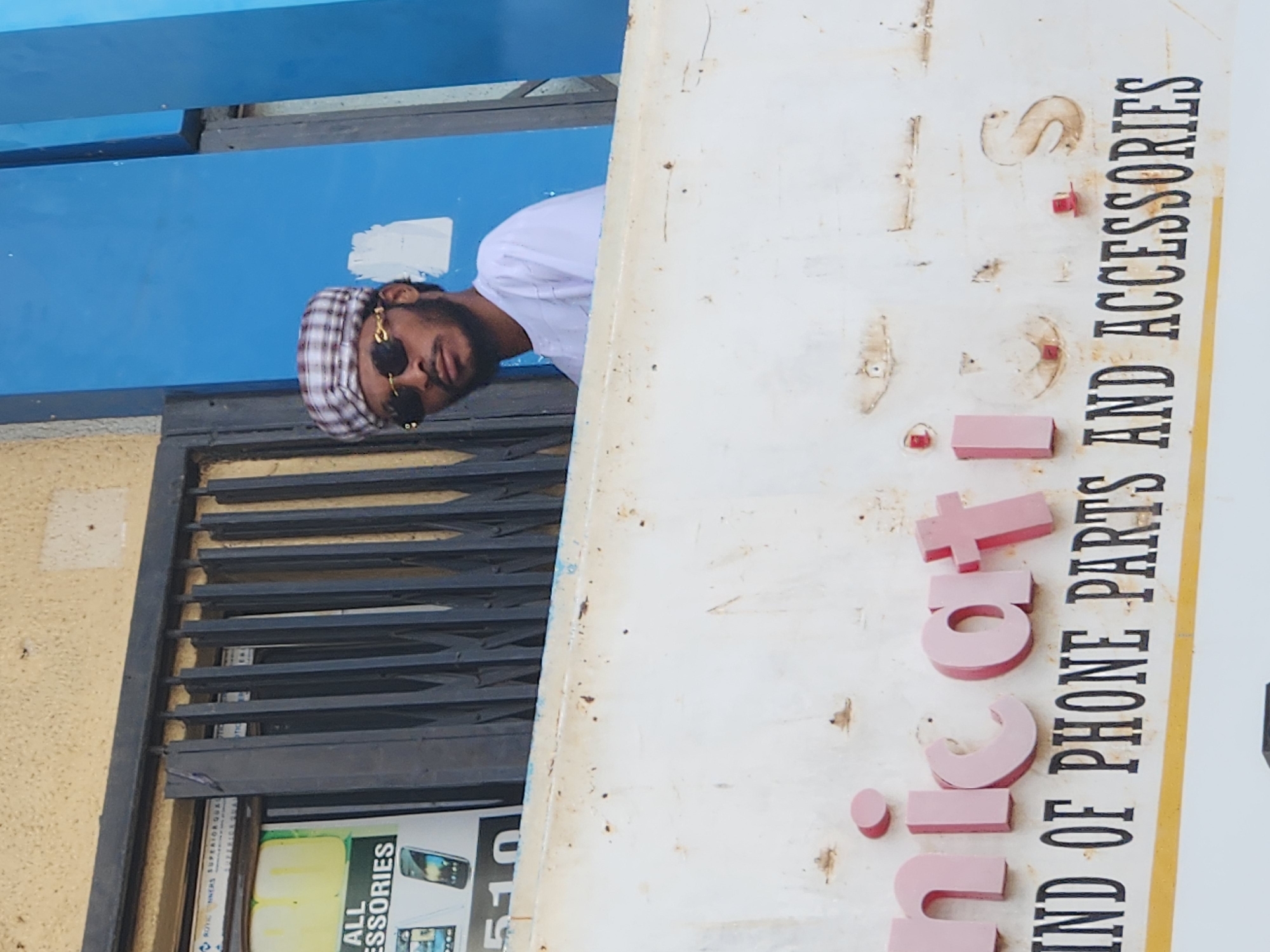 This is a portrait-oriented photograph taken outdoors, likely during the day given the lighting. The main subject is a young Black man, possibly in his late teens or early twenties, looking directly at the camera with a relaxed expression. He is wearing a white t-shirt and a checkered flat cap. He also has on large, dark sunglasses with gold accents.
He is positioned behind a metal grate, which suggests he might be near an entrance or a window of a building. To the right of the man, there is a large, weathered white sign with text. The sign appears to be for a business selling phone parts and accessories. The text on the sign, oriented vertically, reads "IND OF PHONE PARTS AND ACCESORIES". Some of the letters are raised and pink, appearing to spell out "JUICIT." Below this, on a different sign visible through the grate, are images of mobile phones and the word "ACCESSORIES". The background includes a bright blue structure above the man and a textured yellow wall to the left.
The overall scene suggests a commercial area, likely a shopfront in Jos, Nigeria. The weathered nature of the sign might indicate an established business. The man's gaze and attire suggest he is posing for the photograph.
