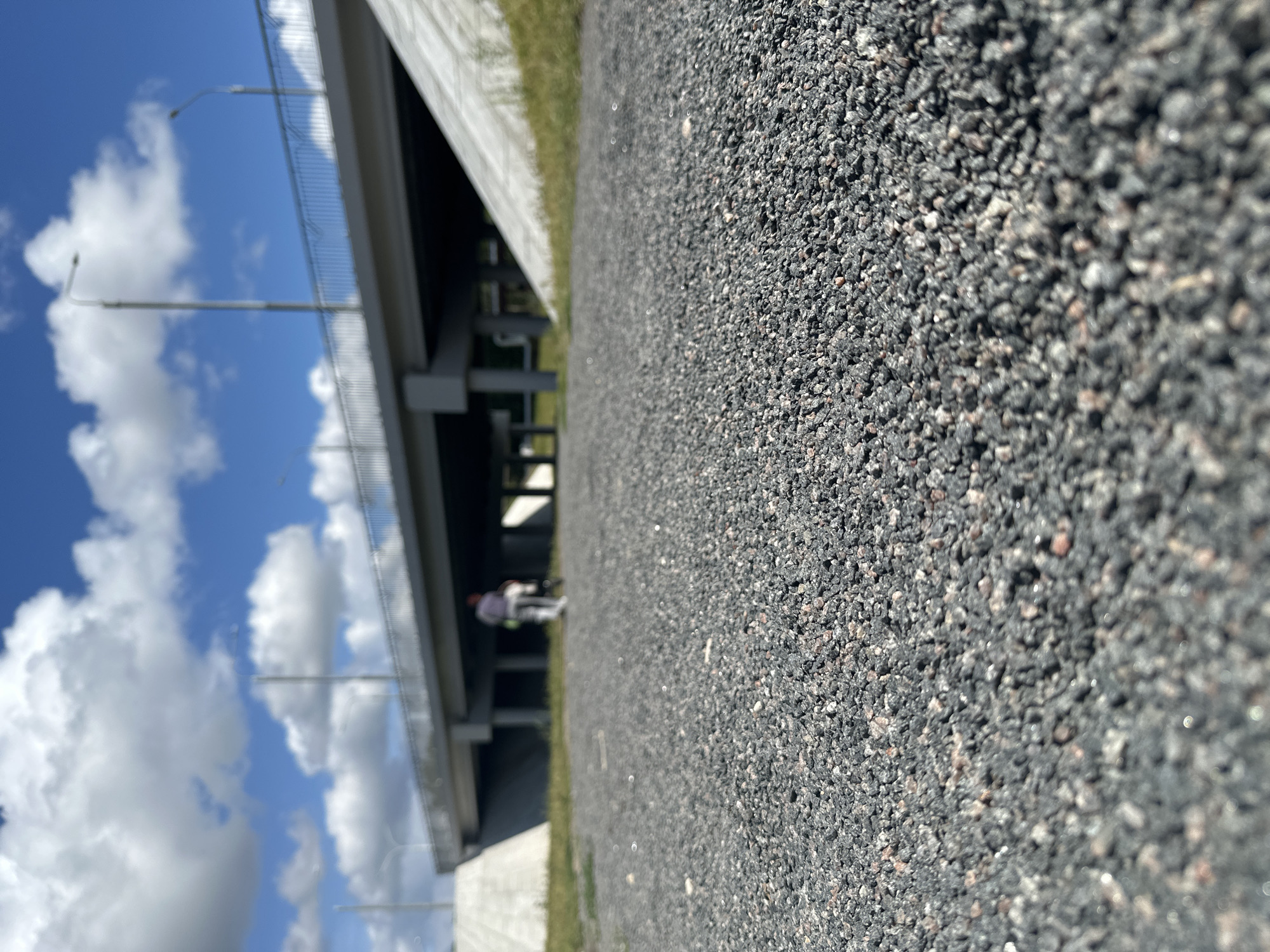 The image depicts a low-angle shot of a gravel path with a blurred background. In the background, a person is seen walking away from the camera, pushing a stroller or cart. Behind the person, a bridge with streetlights and railings stretches across the scene. The sky is blue with scattered white clouds. The environment appears to be an outdoor, possibly park-like setting, with grass visible on the sides of the path and under the bridge. The overall scene suggests a casual, daytime activity. The location is identified as Minsk, Belarus.