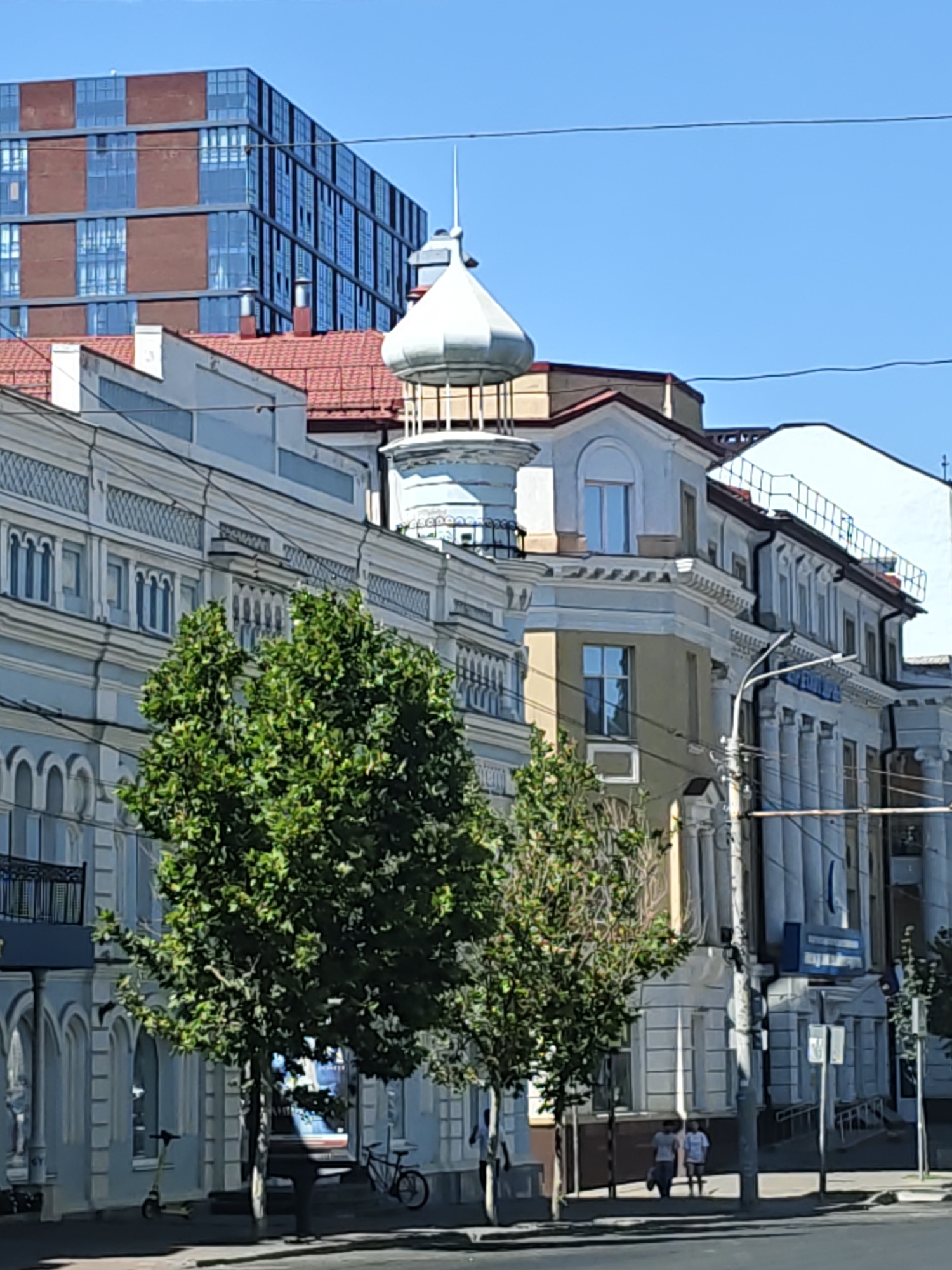This image captures a street scene in Rostov-on-Don, Russia. In the background, a modern multi-story building with a reddish-brown facade and large glass windows dominates the skyline. In the foreground, older architecture is visible. A light blue building with ornate detailing and arched windows stretches across the frame, its facade decorated with decorative patterns and white columns. A distinctive domed structure, possibly a cupola, rises above this building, topped with a slender spire. Two trees with lush green foliage are positioned in front of the older building, partially obscuring its lower levels. A street light and traffic signs are present along the sidewalk, where two individuals are walking. A bicycle and an electric scooter are parked near the sidewalk. The sky is clear and blue, indicating a sunny day.