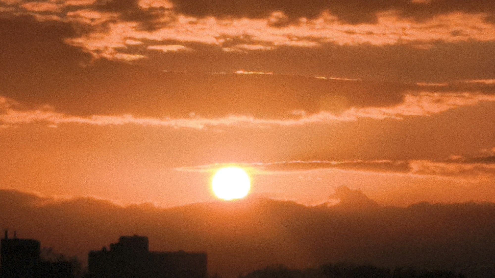 A vibrant sunset paints the sky with shades of orange and yellow, casting a warm glow over the cityscape of Worms, Germany. The sun, a brilliant white orb, is partially obscured by clouds, creating a dramatic effect. Silhouetted against the fiery sky are the outlines of buildings, including what appears to be the distinctive shape of the St. Peter Cathedral in the distance. The lower part of the image is dominated by the dark silhouettes of rooftops and trees, suggesting a densely populated urban area. The overall impression is one of natural beauty and urban life coexisting under a spectacular sky.