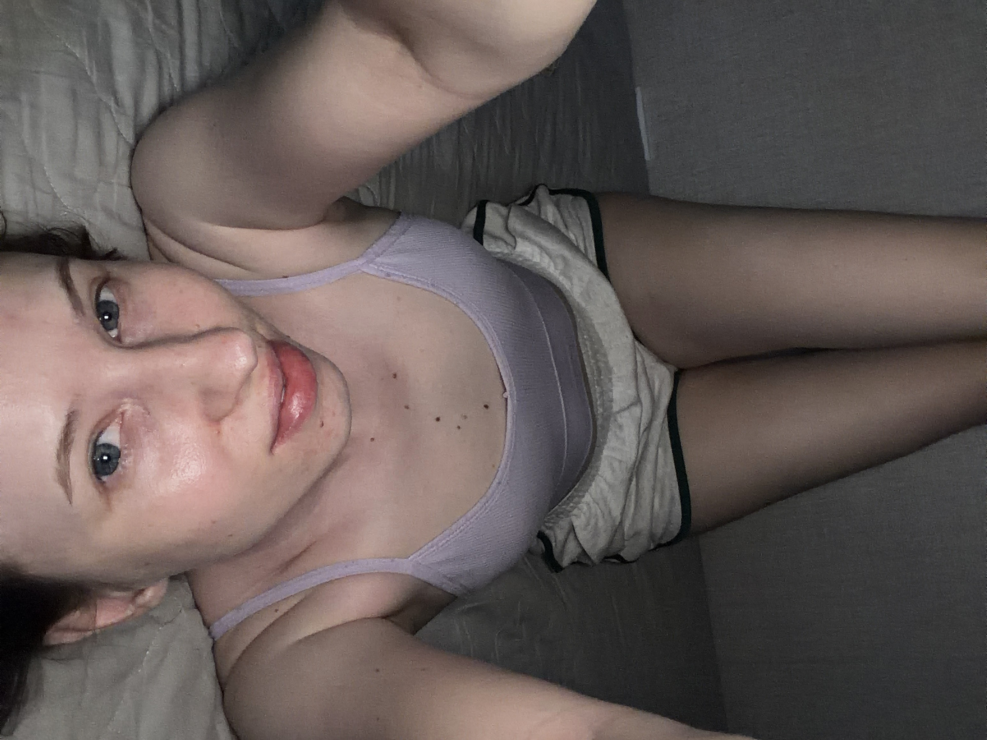 A person is lying down on a bed, with their head turned towards the camera. They are wearing a light purple tank top and light-colored shorts with a dark trim. The person has light skin, blue eyes, and light brown hair. They are looking directly at the camera with a neutral expression. The setting appears to be a bedroom, with a gray textured blanket or comforter visible behind them. The lighting is dim, suggesting it could be nighttime or a room with low light. There is no visible text in the image. The scene is a close-up shot of the person's upper body and face.