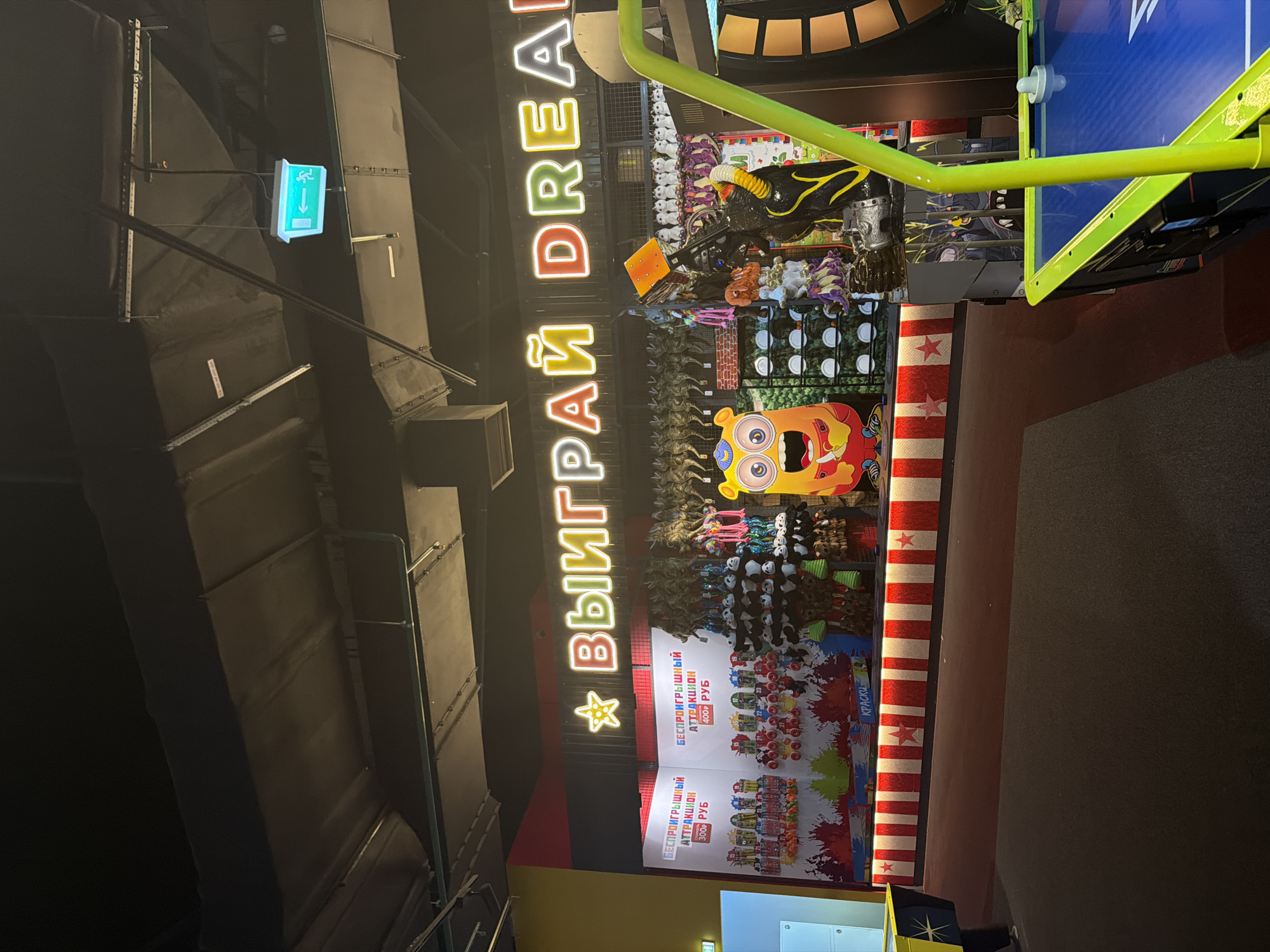 The image shows the interior of an amusement center or arcade. In the background, a brightly lit neon sign with Russian text "ВЫИГРАЙ DREAM" (meaning "Win Dream") is visible against a dark ceiling and industrial ventilation system. Below the sign, a prize counter is set up with rows of stuffed animals and toys displayed on shelves. A large, cartoonish monster mascot with a yellow body and blue accents stands to the left of the counter. The counter itself has a red and white striped facade adorned with red stars. To the right of the counter, there's a portion of an air hockey table. In the foreground, another air hockey table with a blue surface and yellow accents is partially visible. An illuminated green exit sign with an arrow pointing left is mounted on the ceiling. The overall environment is dark, illuminated by the neon signs and the lighting from the arcade machines and displays.