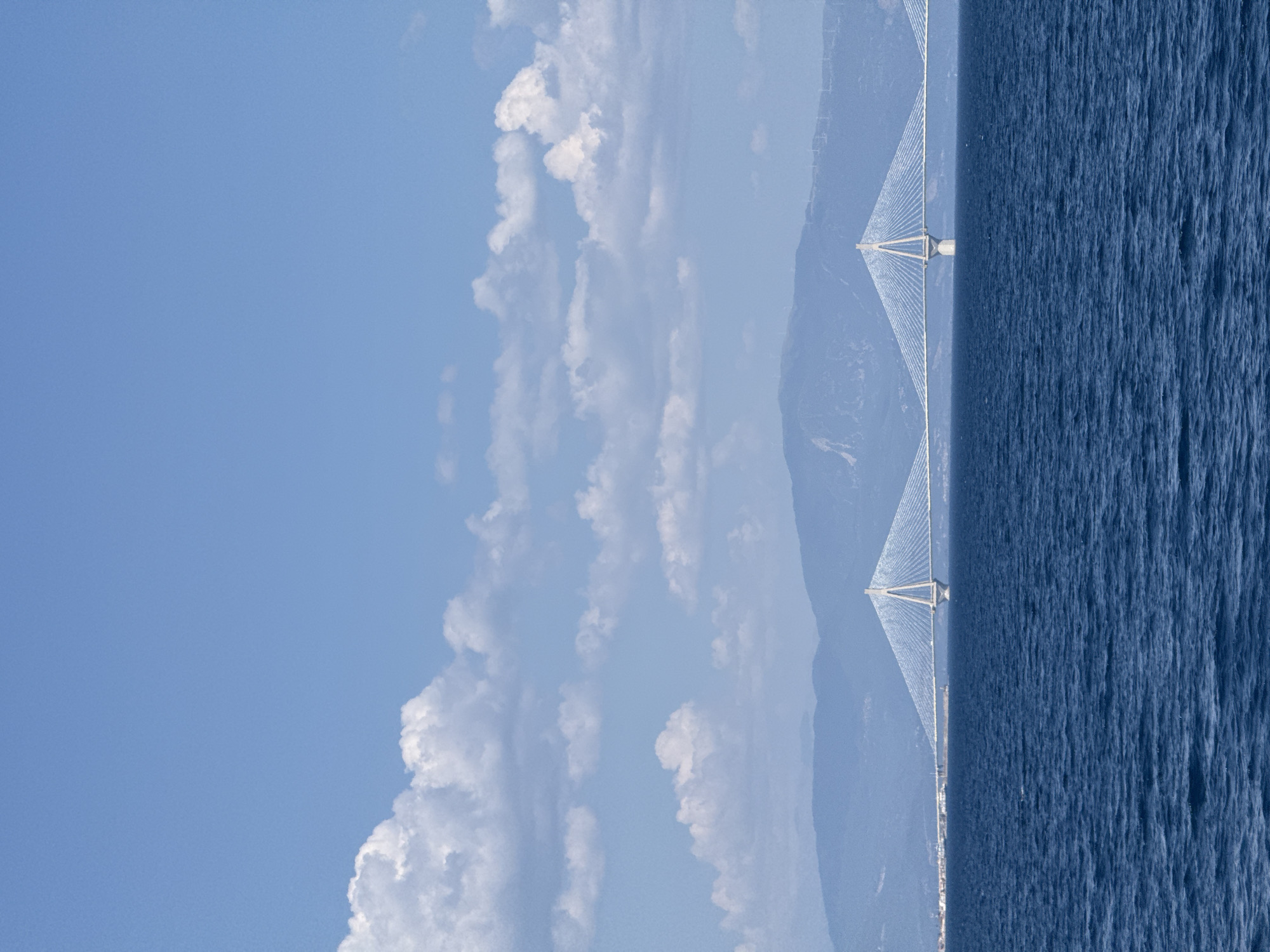This media file depicts a panoramic view of a suspension bridge spanning across a body of water, with a mountainous landscape in the background. The scene is split horizontally, with the sky and clouds occupying the upper portion and the bridge, water, and land filling the lower half.
The sky is a clear, bright blue, punctuated by scattered white, puffy clouds. The sea is a deep, rich blue with a textured surface, indicating gentle waves or ripples. The bridge, a prominent object, is a modern suspension bridge with two tall, white pylons that support numerous cables forming a V-shape. The bridge extends across the frame, connecting the land on either side.
In the distance, hazy blue mountains rise behind the bridge. A distant shoreline with some buildings can be seen along the base of the mountains. The overall atmosphere is serene and majestic, suggesting a calm and pleasant day. The lighting indicates it is daytime, likely mid-morning or afternoon, with the sun illuminating the scene.
The location is identifiable as Patras, Greece, known for its impressive bridges connecting the Peloponnese peninsula to the mainland. The image captures the grandeur of the engineering feat against the natural beauty of the Greek landscape. No people are visible in the image, and there is no visible text.