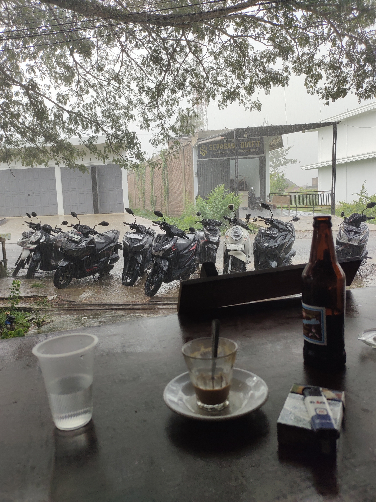 The image depicts a rainy scene in front of a building with the sign "SEPASAN OUTFIT" in Mee, Indonesia. Several motorcycles are parked outside the building. In the foreground, on a table, there is a plastic cup filled with water, a small glass of coffee with a spoon, a pack of cigarettes with a lighter, and a bottle of beer. The overall atmosphere is gloomy due to the rain.