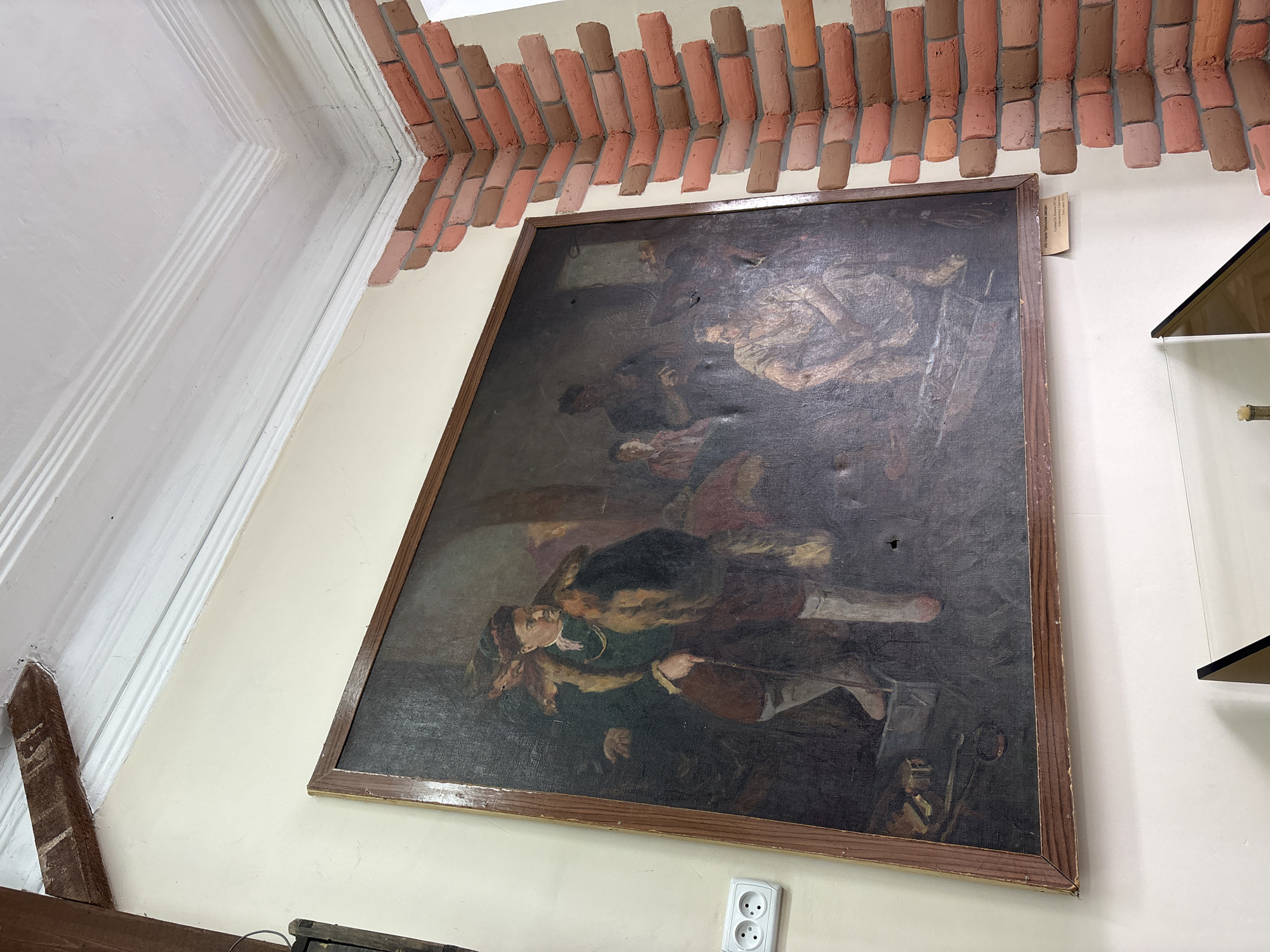 The image shows a large, framed oil painting hanging on a pale yellow wall in what appears to be a museum or gallery. The painting depicts a group of people in a dimly lit, interior setting. The style of the painting is realistic, with a focus on the figures' clothing and expressions.
In the foreground on the left, a man in a fur-trimmed coat and a hat stands with a cane, looking to the right. He is dressed in dark trousers, white stockings, and a green tunic. Behind him, other figures are visible, including a woman in a red garment and a man in the background. In the lower right of the painting, a seated figure in lighter clothing is depicted. The overall atmosphere of the painting is somber and historical.
The painting has visible signs of age, including some damage to the canvas, such as small holes. A small, yellow label is affixed to the bottom edge of the painting's frame, with text that is partially visible.
The wall the painting hangs on is light yellow, and to the right, a corner is decorated with faux red brickwork. Above the painting, a white ceiling with decorative molding is visible, and a wooden beam runs diagonally across the top left of the frame. A white double electrical outlet is mounted on the wall to the left of the painting. The environment suggests an indoor exhibition space.
The painting is located in the Suksun Museum of Local Lore in Suksun, Russia. The label on the painting indicates it is titled "For the Sconces," created in 1957 by artist V.B. Tikhonov.