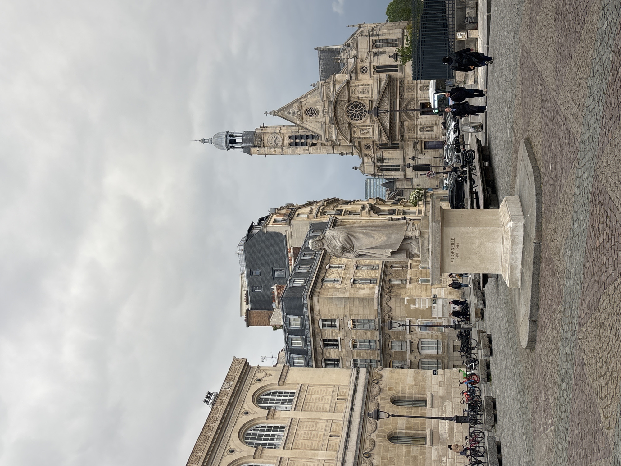 The image shows a street scene in Paris, France, featuring the Saint-Gervais-Saint-Protais church in the background. The church's facade and bell tower are visible, showcasing its architectural details. In the foreground, there is a statue on a pedestal. Parked bicycles are seen near the statue. People are walking and standing nearby. The street is paved with cobblestones. Buildings with Parisian architecture line the street. The sky is overcast. The atmosphere appears to be a typical day in the city.