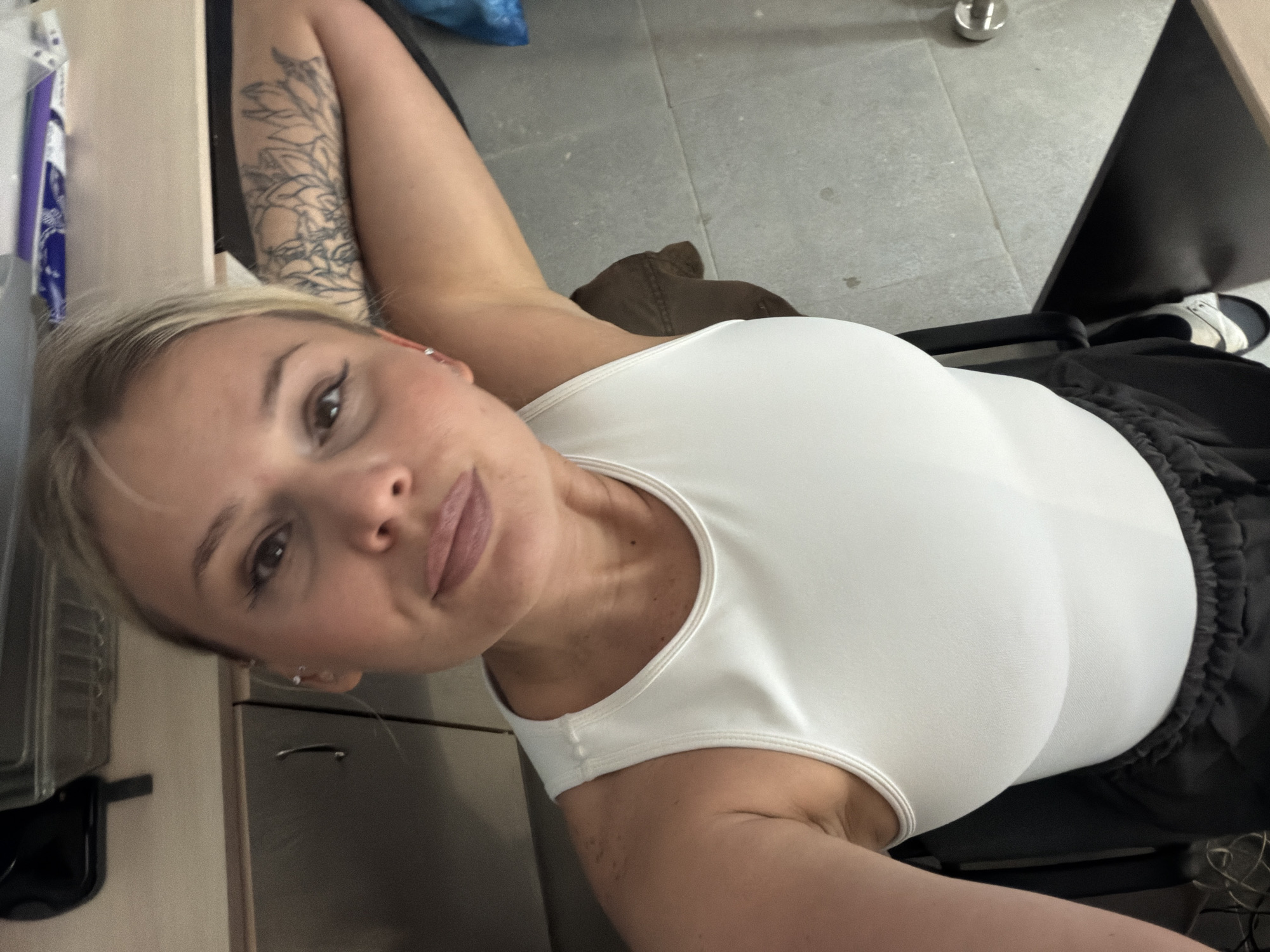 A fair-skinned woman with blonde hair is seated in a chair, wearing a white top and black bottoms. Her left arm, adorned with a tattoo, is raised behind her head. The setting appears to be indoors with a gray floor, and the background includes a countertop with various objects. There is no indication of the city of Sarapul, Russia, in the image.