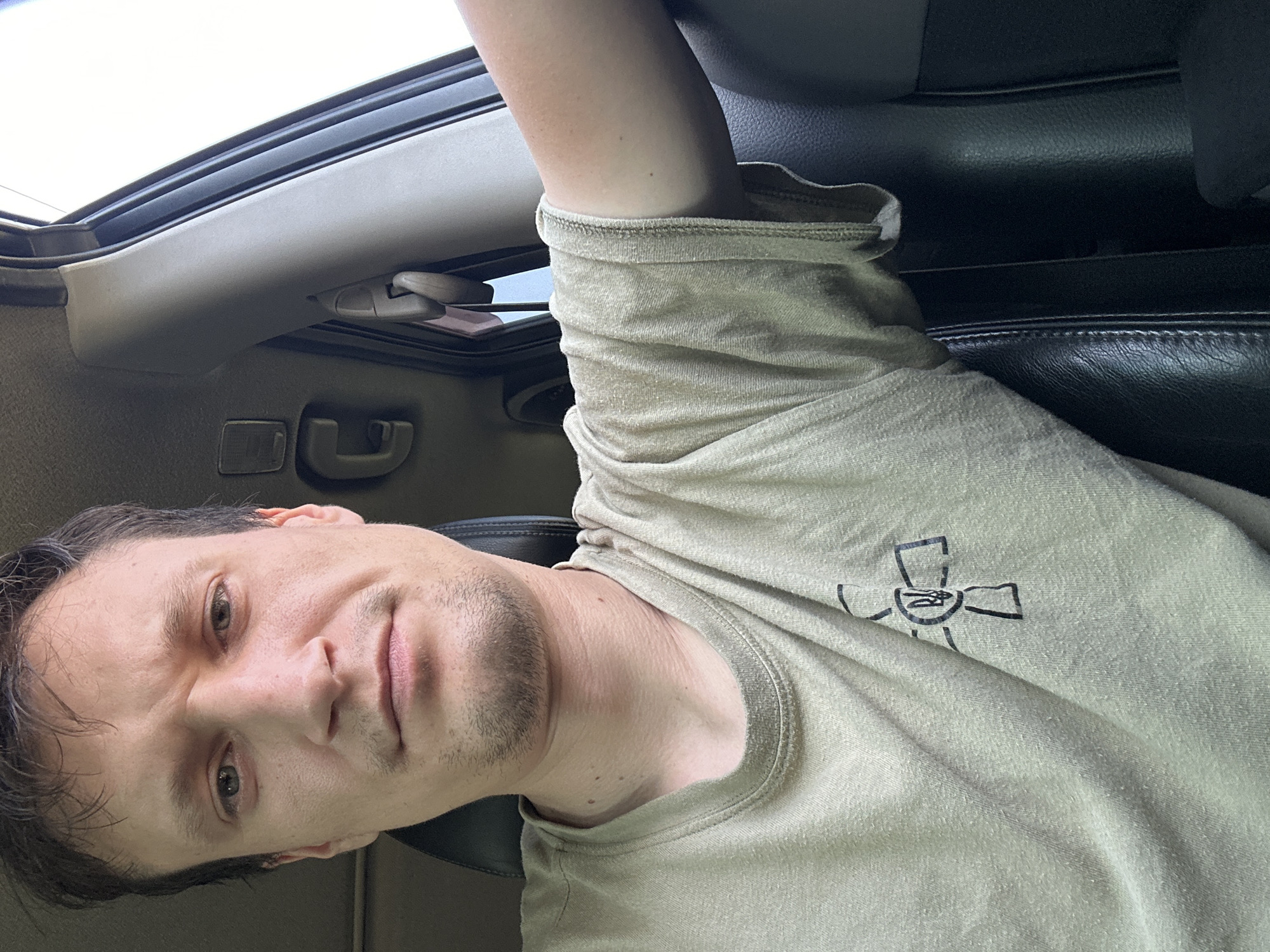 A man with brown hair and stubble is sitting in the driver's seat of a car. He is wearing a light brown t-shirt with a symbol printed on the left side of his chest. The symbol appears to be a cross with a trident inside. The interior of the car is visible, including the black leather seat, a grey pillar, and a black door handle. The background is blurry, but appears to be outdoors with bright light. The image appears to be taken by the man himself, suggesting a selfie. The location is Makiv, Ukraine.