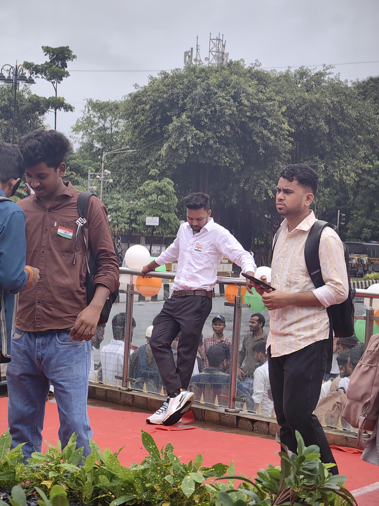 In Mumbai, India, on a cloudy day, a group of young men stand on a red platform, possibly during a celebration or event. A young man in a brown shirt with a small Indian flag pinned to his pocket smiles as he looks down. To his right, another young man in a white shirt leans on a railing, his head tilted back. Further to the right, a young man in a patterned shirt holds a mobile phone and looks to the side with a serious expression. In the background, trees, traffic, and other people are visible, suggesting an urban setting. The platform is bordered by a glass railing adorned with orange and white balloons.