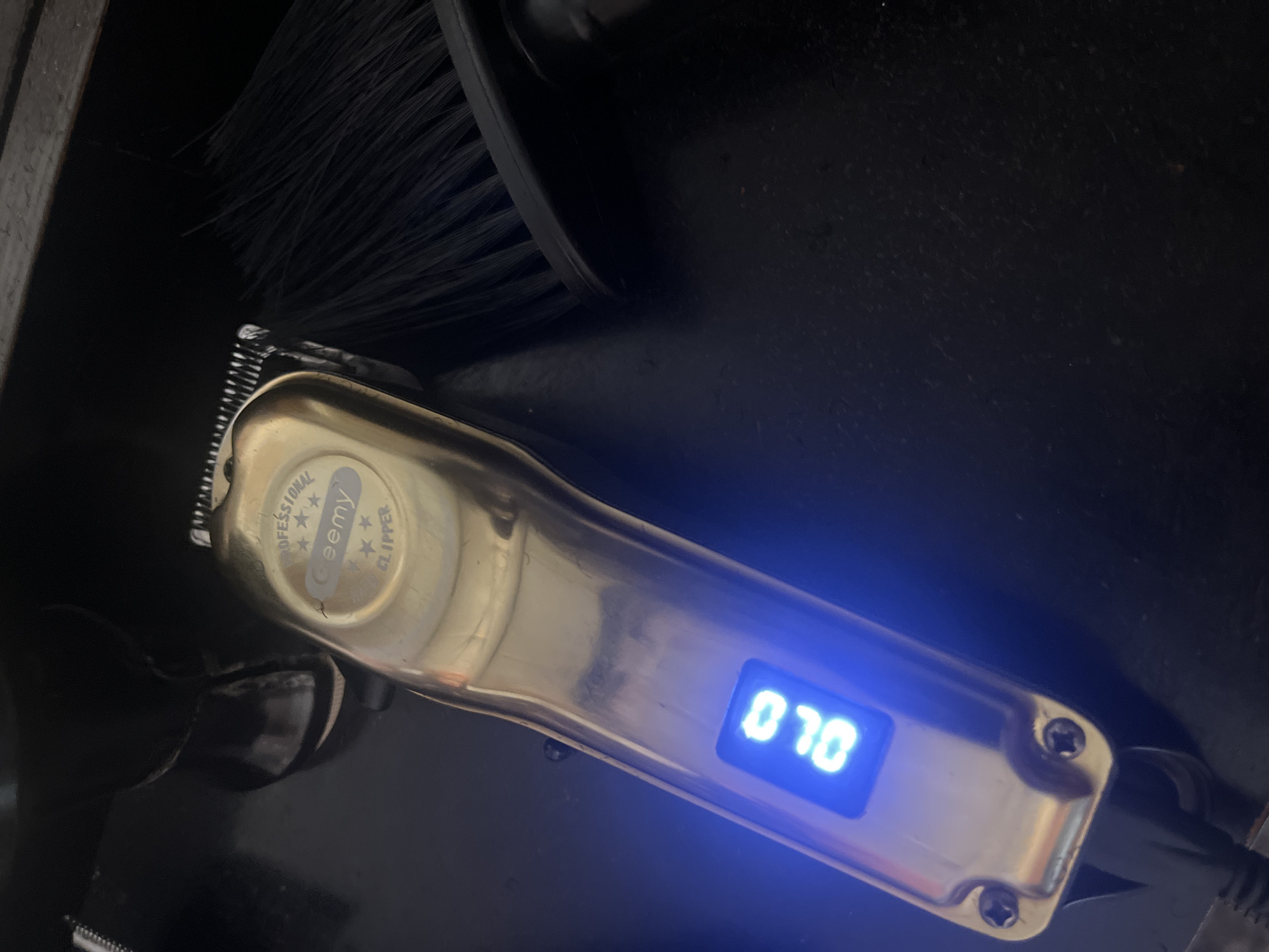 A gold-colored electric hair clipper is positioned on a dark surface near a black brush. The clipper has an illuminated digital display showing "070." The clipper's head is near the brush. The scene is darkly lit. No people are visible. The environment suggests a grooming or barbering context. The scene's location is not evident from the image.