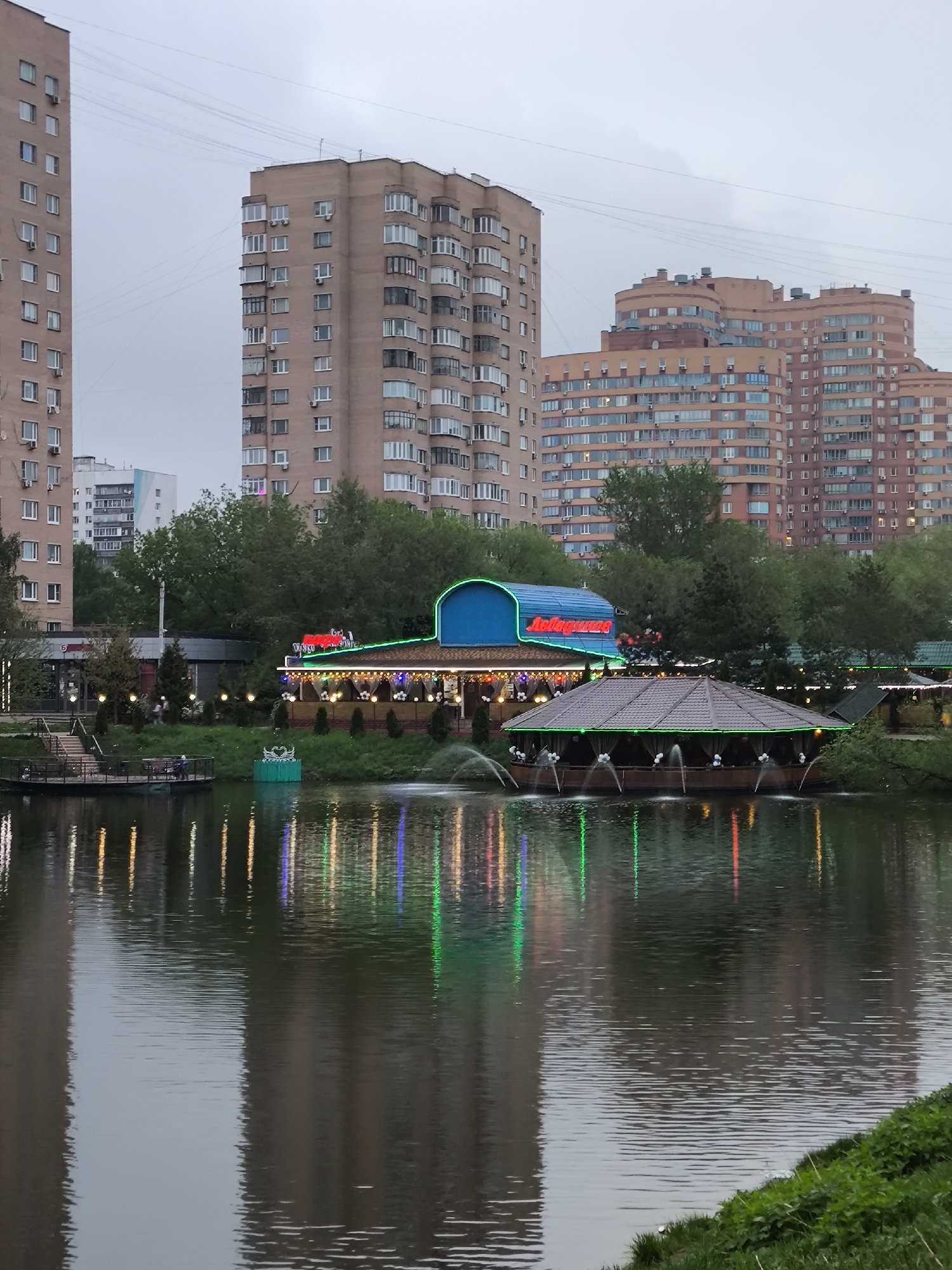 The image depicts a view across a body of water, likely a lake or large pond, in what appears to be an urban or suburban setting. The photograph seems to be taken in the evening, as evidenced by the muted light and the illumination of various structures.
In the foreground, there is water with distinct ripples on its surface. Reflections of the surrounding structures and lights are visible in the water, creating vertical streaks of various colors. Green vegetation lines the edge of the water in the bottom right corner.
Across the water, there are two prominent structures. The first is a pavilion or restaurant, identifiable by a dark, tiled roof, supported by columns. It is illuminated by green neon lights. Water fountains are seen shooting up from the perimeter of this structure, further adding to its distinctive appearance. The second structure is a building with a bright blue roof. A sign in a foreign language, illuminated with red neon, is visible above the entrance.
Behind the pavilion, there are several multi-story residential buildings. These buildings are constructed of light brown brick or concrete. Trees are scattered between the water and the buildings.
The overall atmosphere is calm and subdued. The interplay of light and shadow, along with the reflections in the water, creates a tranquil urban scene.
Based on the location of the image, it is likely that this is located in the city of Khimki, Russia.