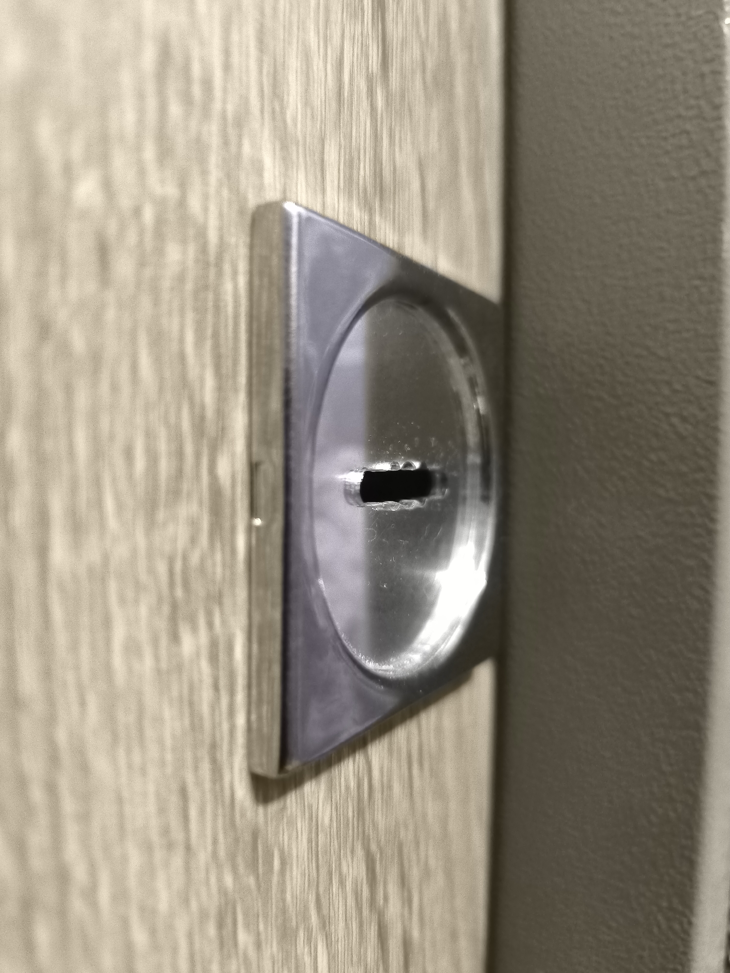 The image shows a close-up, eye-level view of a metallic escutcheon plate on a wooden surface. The escutcheon is square with rounded corners and features a circular recess containing a horizontal keyhole slot. The surface of the escutcheon is reflective, showing smudges and glints of light. The wooden surface appears to be a light brown with a prominent wood grain pattern, likely a door or furniture. To the right of the escutcheon, a textured, light grey surface is visible, suggesting it's the edge of another object or a wall. The lighting suggests an indoor environment, possibly with artificial light casting shadows and highlights. There are no people or active events depicted. The image is a still shot focusing on the hardware detail. The location in Horishni Plavni, Ukraine, is not discernible from the visual information in the image itself.