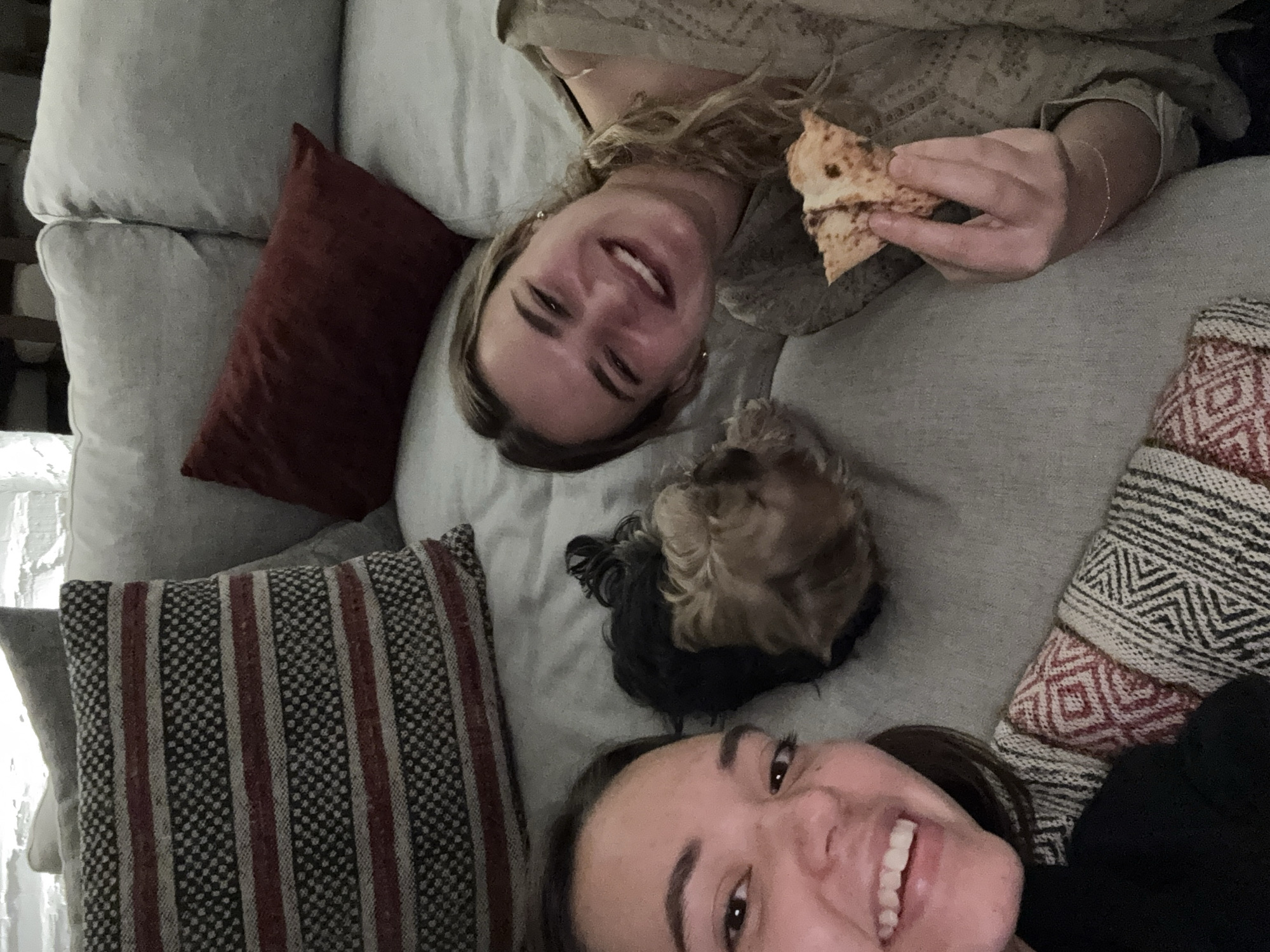 Two women and a small dog are reclining on a light gray couch with several pillows. One woman is blonde and holding a piece of food. The other woman has dark hair and is smiling. The couch has a red rectangular pillow, a striped pillow and a patterned pillow. The location is indeterminable.