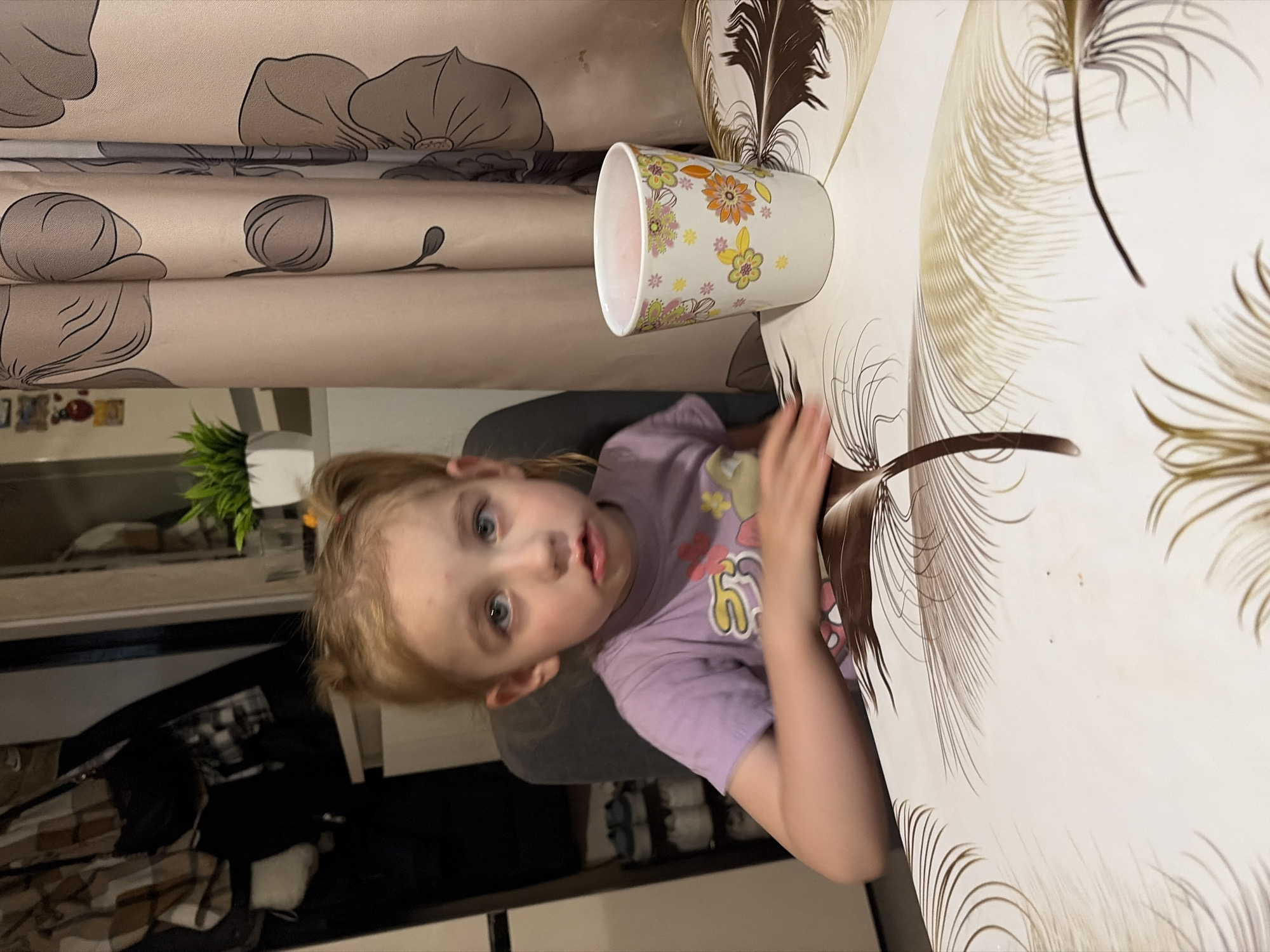 A young girl with blonde hair in pigtails, wearing a light purple t-shirt, sits at a table in what appears to be a dining area. She has blue eyes and a neutral expression. In front of her is a white mug with a floral pattern. The table is covered with a white tablecloth featuring a brown and tan feather design. Behind the girl, to her right, is a beige curtain with a large floral pattern in shades of brown. To her left, a closet or wardrobe is visible, with clothing hanging on hooks. A small potted plant sits on a shelf in the background. The scene appears to be indoors, in a residential setting in Tambov, Russia.