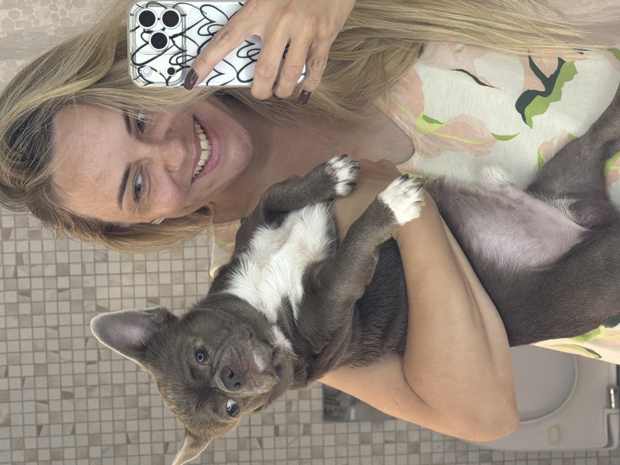 In a bathroom with tiled walls, a fair-skinned woman with long blonde hair smiles as she holds a small, grey-and-white dog. The woman is taking a selfie with her phone, which has a clear case decorated with black heart outlines. The dog, held in her arms, is looking towards the camera with its ears perked up. The dog's fur is short and dense, with white markings on its chest and paws. A floral-patterned shirt is visible on the woman. The overall scene is candid and intimate, capturing a moment between the woman and her pet. There is no specific information to indicate this scene is located in Votkinsk, Russia.