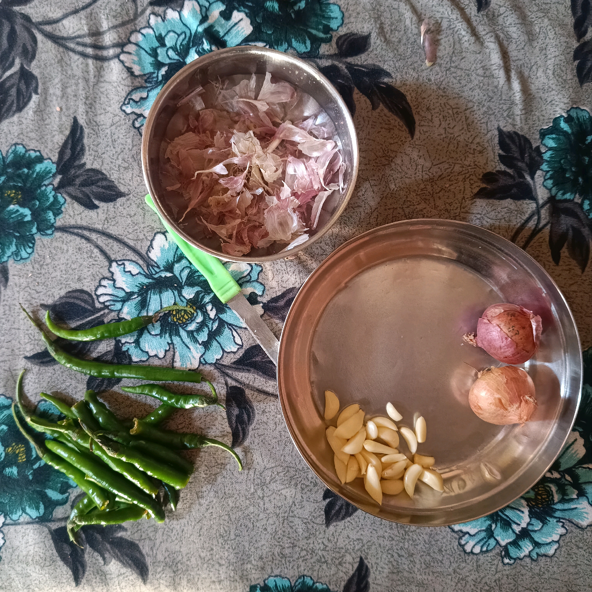 A close-up, overhead shot shows a collection of ingredients for cooking on a floral patterned cloth. In the upper left, a silver bowl contains a pile of thinly sliced garlic. To the right, a silver plate holds two whole, small red onions and a pile of peeled garlic cloves. In the lower left, a bundle of green chili peppers is arranged on the cloth. A small green-handled knife is positioned between the bowl of garlic slices and the chili peppers. The cloth has a grey background with blue and dark green floral designs. The scene appears to be indoors, on a flat surface.