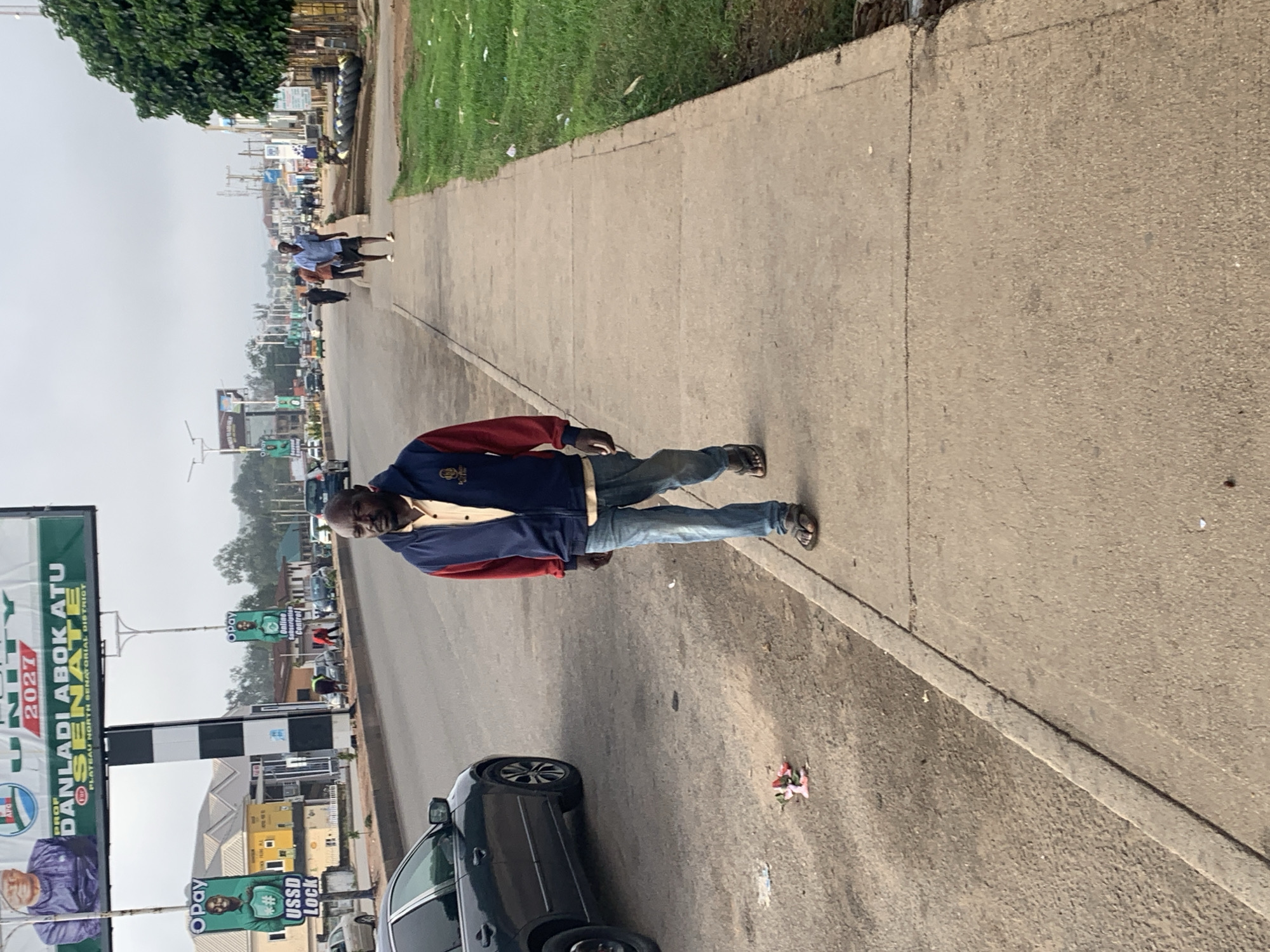 The image shows a street scene in Gura Top, Nigeria. A man in a blue and red jacket and jeans walks on a concrete sidewalk. To his left, a dark-colored sedan is parked on the road. In the background, there are several billboards and buildings lining the street. Traffic is visible on the road, including cars and a yellow minivan. The sky is overcast.