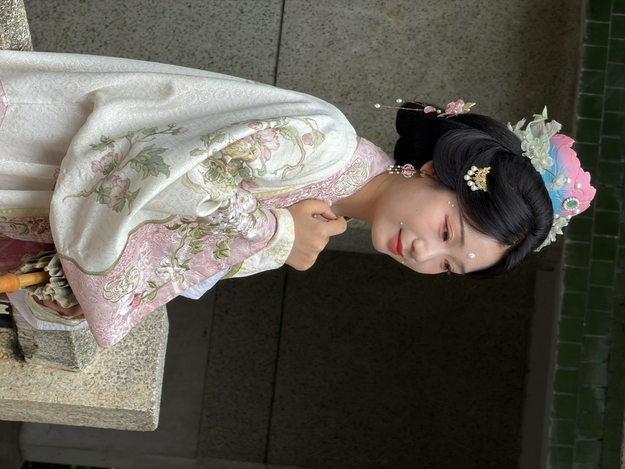 A young East Asian woman stands on a stone structure, facing right in profile. She wears a traditional Hanfu-style garment with a white outer layer featuring a floral pattern in pink and green. Underneath, a pink garment with similar embroidery is visible. Her hair is styled in an elaborate updo, adorned with pearl-studded hairpins, delicate flower ornaments, and a colorful headdress featuring pink, blue, and a green gem. She also wears pearl earrings and has decorative white dots on her face, including one on her forehead. Her left hand is raised to her chest, and she holds a folded fan with a wooden handle. The background consists of a textured stone wall, a dark opening, and a green tiled wall above. The location is identified as Yuzhong District, China.