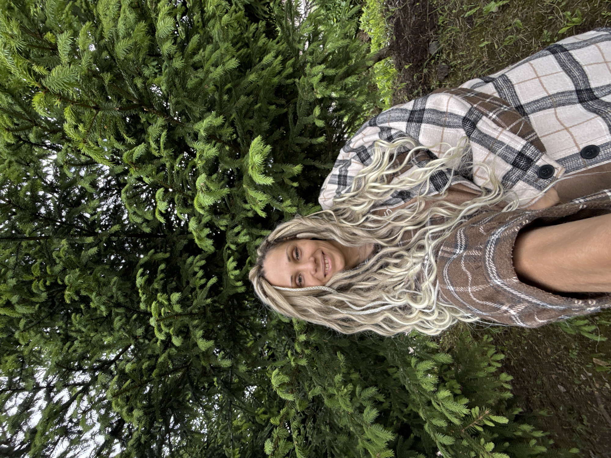 A woman with blonde dreadlocks is smiling at the camera, positioned against a backdrop of a dense evergreen tree. She is wearing a brown and beige plaid jacket. The location is likely Rusanovo, Russia, as specified in the prompt, though no features in the image confirm it. The scene is outdoors, with hints of other vegetation and soil visible around the tree and the woman. The woman appears to be interacting with the camera, likely posing for a photograph.