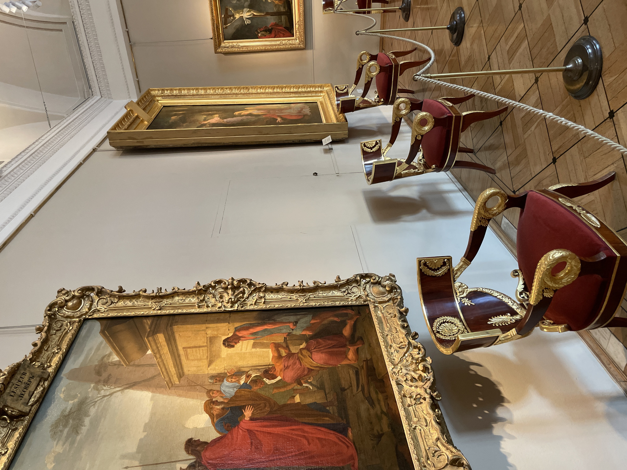 The image shows an interior of a museum in Saint Petersburg, Russia. Three paintings hang on the wall, each with ornate gold frames. The largest painting in the foreground depicts a crowd of figures in biblical attire, rendered in dark, rich colors. Further back, two additional paintings are visible, one depicting a nude figure and the other a crucifixion scene. A series of decorative red and gold chairs line the floor, separated from the paintings by a white rope barrier and gold poles. The floor is made of parquet wood. The walls are a muted beige color.