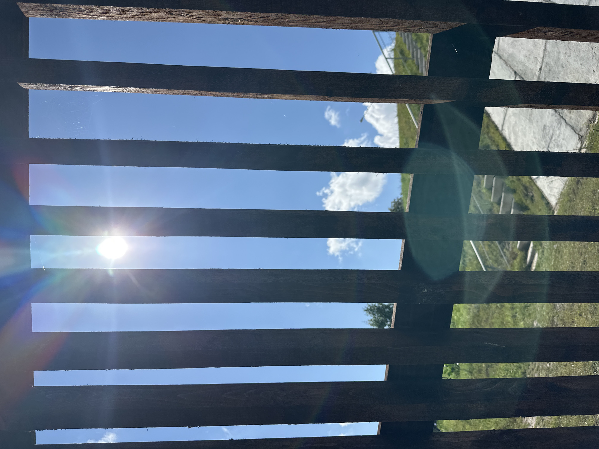 The image shows a view through vertical wooden slats against a bright blue sky with some white clouds. The sun is visible, creating lens flare and a rainbow effect. In the background, there is a grassy incline with concrete steps and a railing, suggesting an outdoor public space. The scene appears to be a sunny day. The location is identified as Minsk, Belarus.