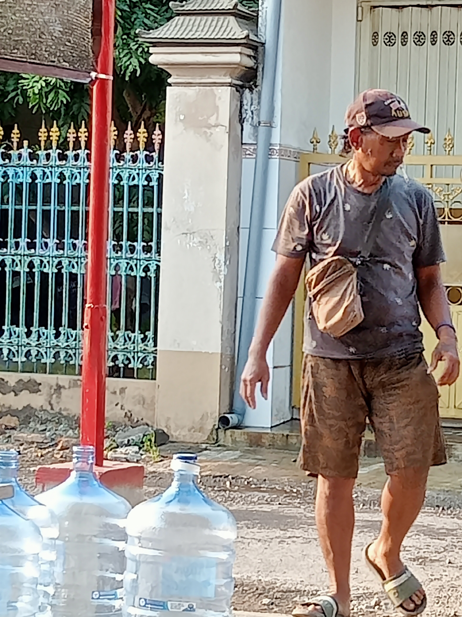 A man is walking in front of a row of large water bottles in the city of Simo, Indonesia. He is wearing a brown baseball cap, a dark grey t-shirt with a pattern, and brown camouflage shorts. A light brown satchel is slung across his chest. The man is wearing light brown sandals on his feet. In the background, a teal metal fence with gold accents is visible, as well as a white wall with ornate carvings. The ground is dirt and gravel.
