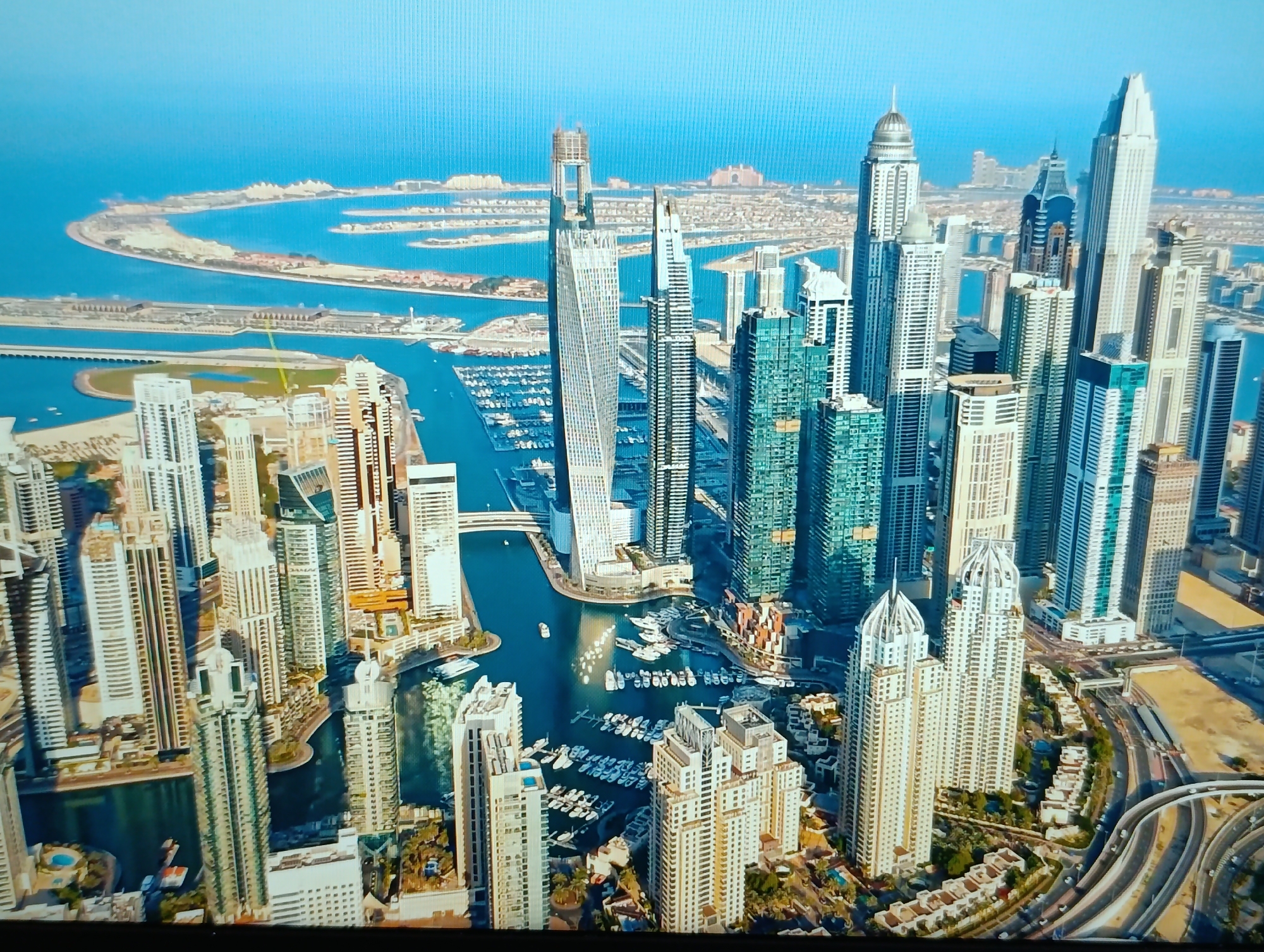 This is an aerial view of a modern cityscape dominated by numerous skyscrapers and a body of water with a marina. The skyscrapers are of varying heights and architectural styles, with many featuring sleek, reflective facades. The marina is filled with numerous boats and yachts. In the background, a distinctive palm-shaped island formation is visible, suggesting the location is likely Dubai, United Arab Emirates, famous for its man-made islands. The water is a vibrant blue, and the sky is a clear, bright blue. There are no people visible in the image. The image is a static aerial photograph without any discernible actions or interactions.
The request to include the location in the city of Dubăsari town council, Moldova, is not consistent with the visual evidence in the image. The scene depicted, particularly the palm-shaped islands and the density of modern skyscrapers, is characteristic of Dubai, United Arab Emirates, not Dubăsari, Moldova. Dubăsari is a city in Moldova that does not feature such extensive modern skyscraper development or artificial island formations. Therefore, it is not possible to accurately describe the image as being in Dubăsari, Moldova.