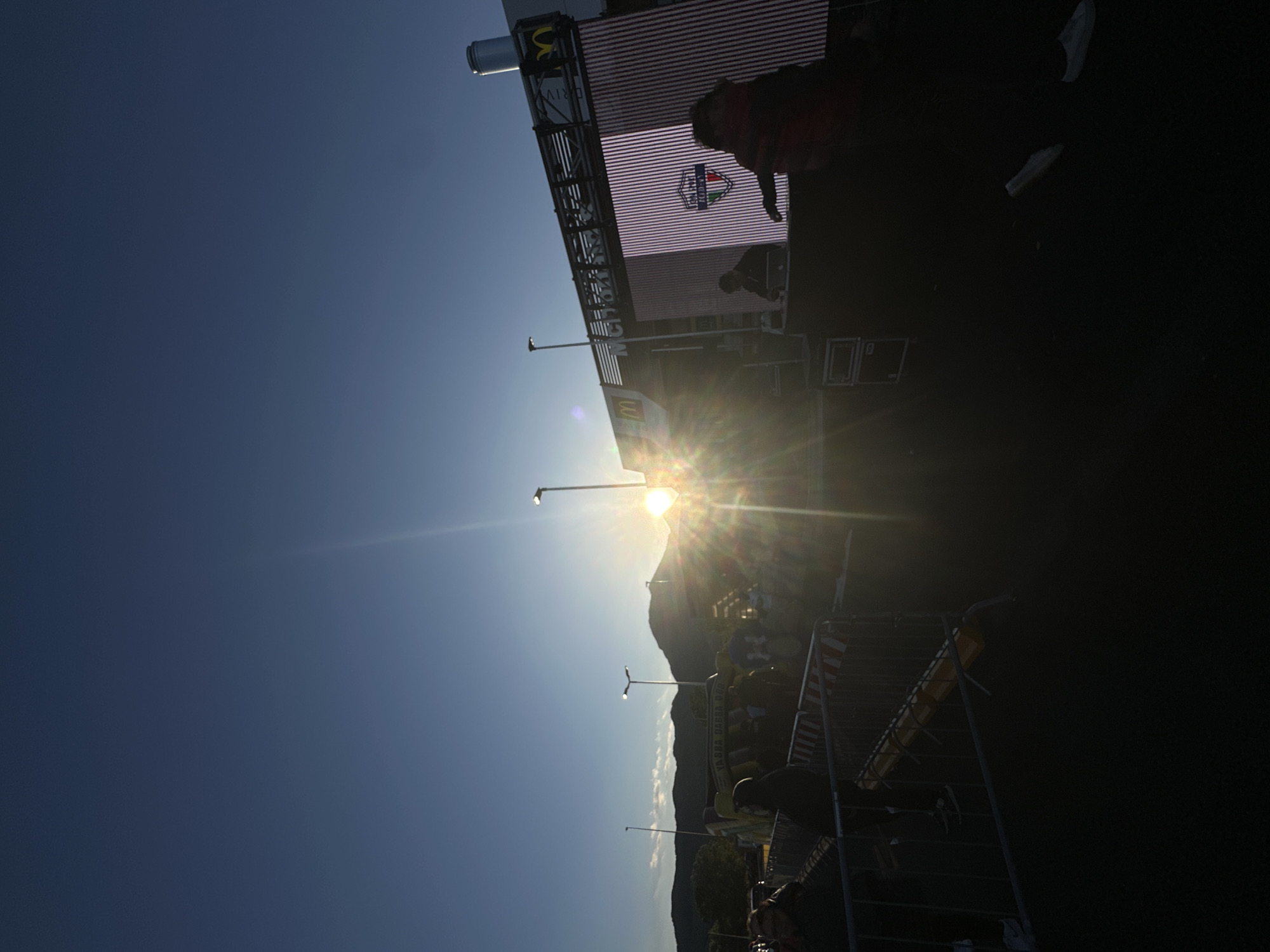 This landscape-oriented image, rotated 90 degrees counter-clockwise in the provided view, captures an outdoor event scene bathed in the dramatic light of either sunrise or sunset. The sun is a powerful focal point, low on the horizon, creating a prominent lens flare and casting long, dark silhouettes across the scene. The sky transitions from a deep, clear blue at the top to a brilliant yellow-orange gradient around the sun.
In the mid-ground, a large stage or event structure is visible, featuring a wide LED screen. On this screen, a crest-like logo is displayed, potentially for a local event or organization, with some text partially legible, possibly referencing "VENAFRO." A McDonald's 'M' logo is also clearly visible on the upper part of the structure. Silhouetted figures of people are present on and around the stage, including one distinct person in front of the screen.
Several streetlights or event lighting poles extend upwards from the mid-ground, their lights appearing as bright points against the sky. In the background, the dark outline of mountains completes the horizon, indicative of a hilly or mountainous location like Venafro, Italy. The foreground is marked by dark metal crowd control barriers, behind which additional silhouetted individuals are visible. Faint text such as "YASSA DANZA 903" appears on one of these barriers. The scene suggests an event either beginning or concluding, with the strong backlighting creating an atmospheric and somewhat mysterious ambiance.
