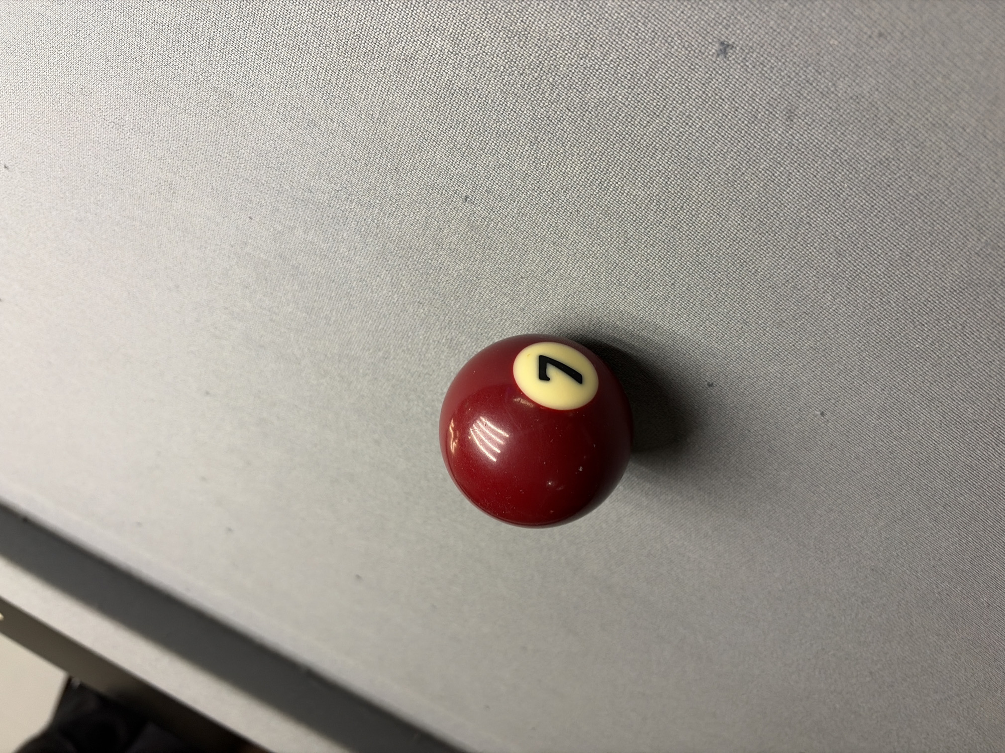 A maroon billiard ball, numbered "7" in white within a cream circle, rests on a light gray, textured surface. The surface may be a billiard table felt. The lighting is diffuse, casting soft shadows around the ball. The bottom left corner of the frame includes a portion of a dark-colored, possibly metallic, edge. There is no information within the image to suggest a location in Budva, Montenegro.