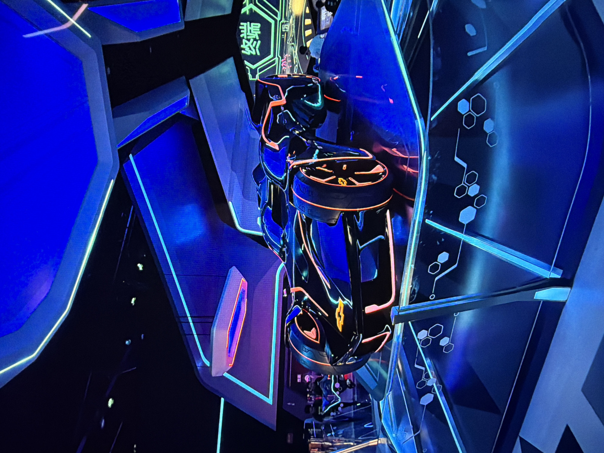 The image shows a futuristic ride vehicle suspended within a dark environment illuminated with neon blue and orange lights. The vehicle is black with orange accents. Geometric shapes and lines, along with Chinese characters, are also illuminated in green neon in the background. The overall scene suggests a Tron-like or science fiction themed attraction. The specific location within the city of Medan, Indonesia is not discernible from the image.