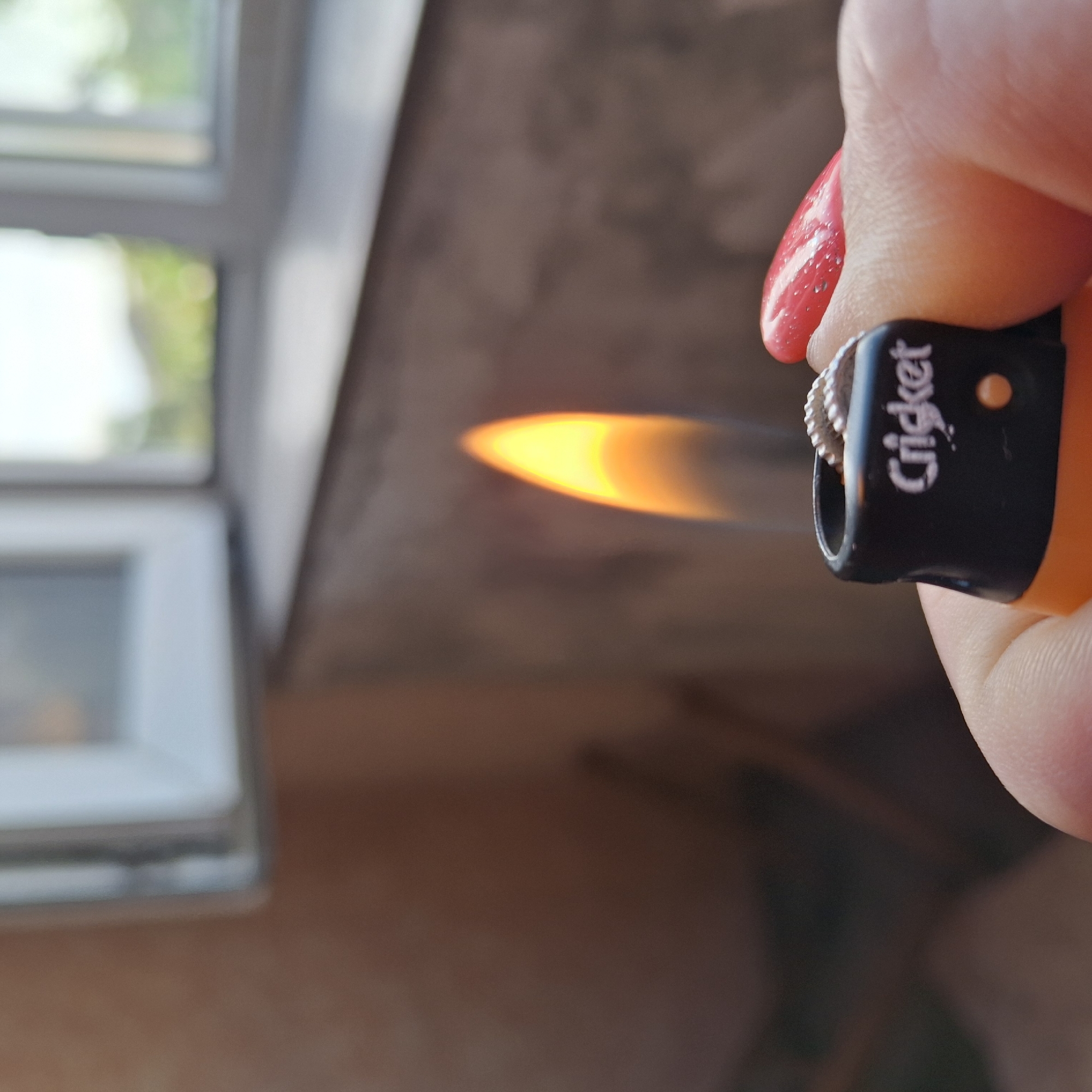 A close-up, slightly blurred image shows a person's hand holding an orange and black "Cricket" brand lighter with a lit flame. The flame is bright yellow and orange. The person's fingernail is painted a bright red. In the background, there is a windowsill and a window with greenery visible outside. The lighting suggests it is daytime.