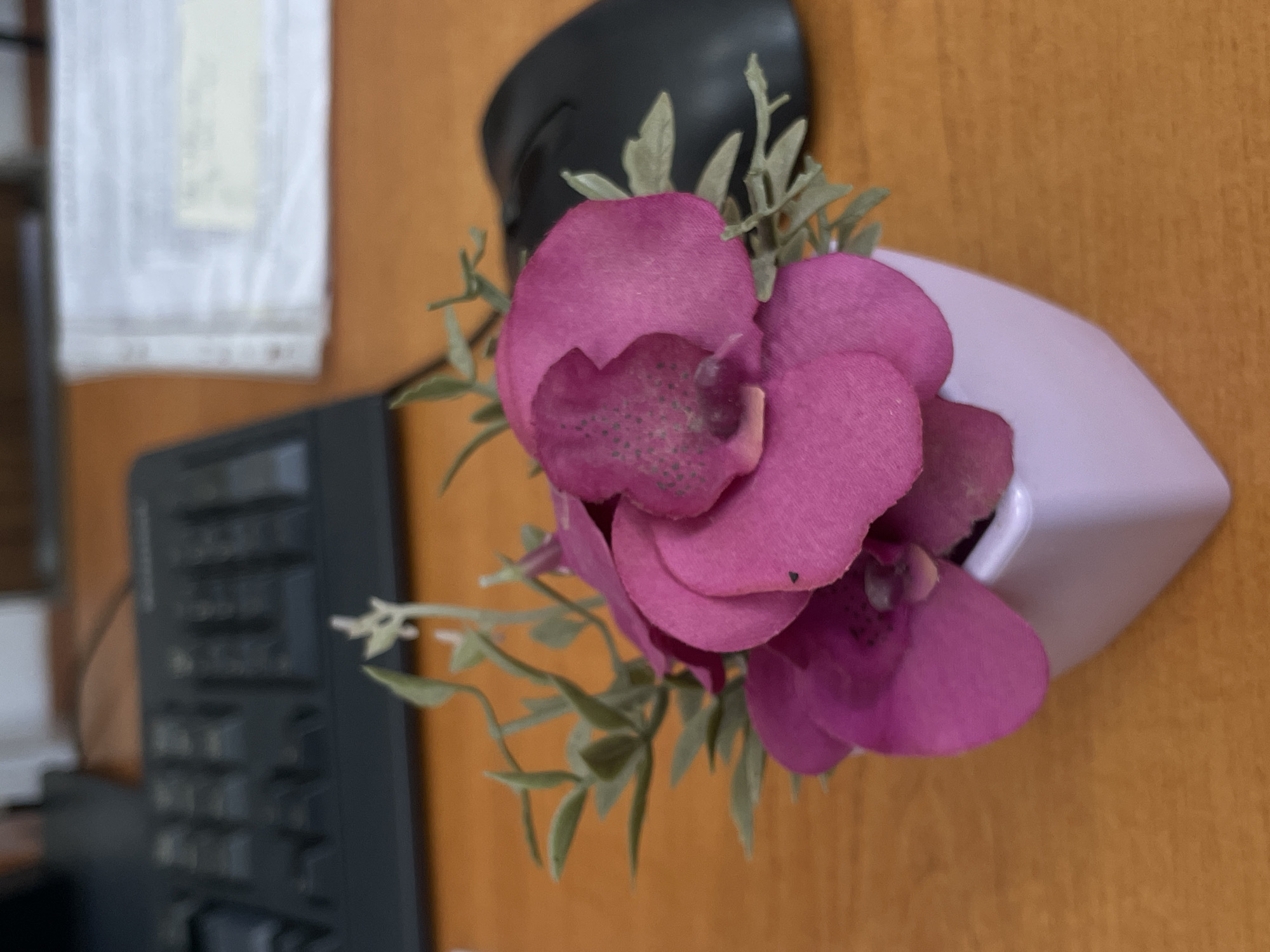 The image depicts a close-up of a small bouquet of artificial pink orchids in a white, geometric vase. The arrangement sits on a light brown, possibly wooden surface, which appears to be a desk or table. To the left of the flowers, a portion of a black keyboard is visible. In the upper left corner, there's a blurred white object that could be a document or decoration hanging on a wall.
The image provides no information about the location being Sungurlare, Bulgaria, or any people, actions, or interactions.