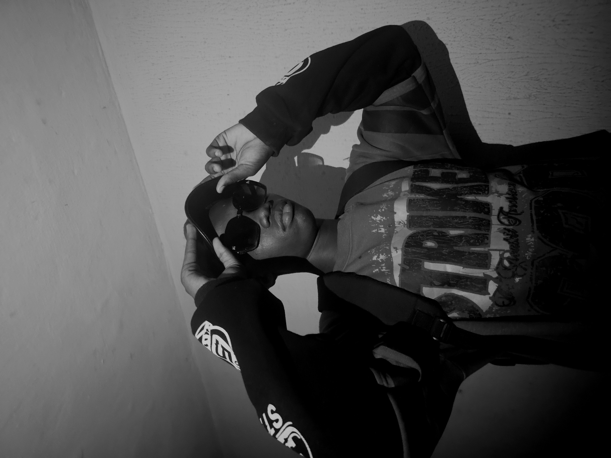 A black and white image depicts a person wearing sunglasses and a patterned sweatshirt. The person appears to be holding the sunglasses with their hand. The background consists of a plain wall, suggesting an indoor setting. The image is tilted, causing the subject and the background to appear at an angle. There is no information about this photo having been taken in Nkwelle, Nigeria.