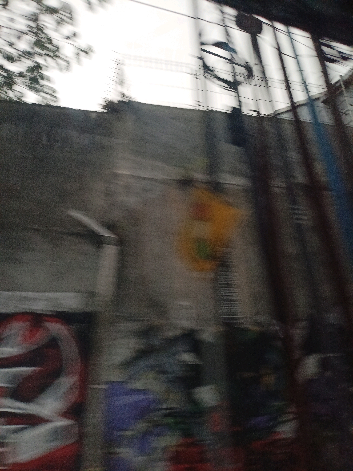 The image captures a blurred scene with a concrete wall and various objects that appear to be in motion. There's a large, somewhat abstract red and white circular graffiti tag on the left side of the image. Adjacent to it, a patch of purple graffiti is visible. Above this, on the wall, a yellow object with some green markings is present. Vertical lines, possibly poles or bars, run along the right side of the image, some appearing as dark, weathered metal and others with a bluish tint. Overhead, a tangle of electrical wires and poles are visible against a bright, possibly overcast sky. The overall impression is that of a quick, possibly vehicle-mounted, photograph taken in an urban environment, given the presence of graffiti and utility infrastructure. While no specific landmarks in Magelang, Indonesia are identifiable due to the blur and lack of context, the elements suggest a typical street scene in a built-up area.