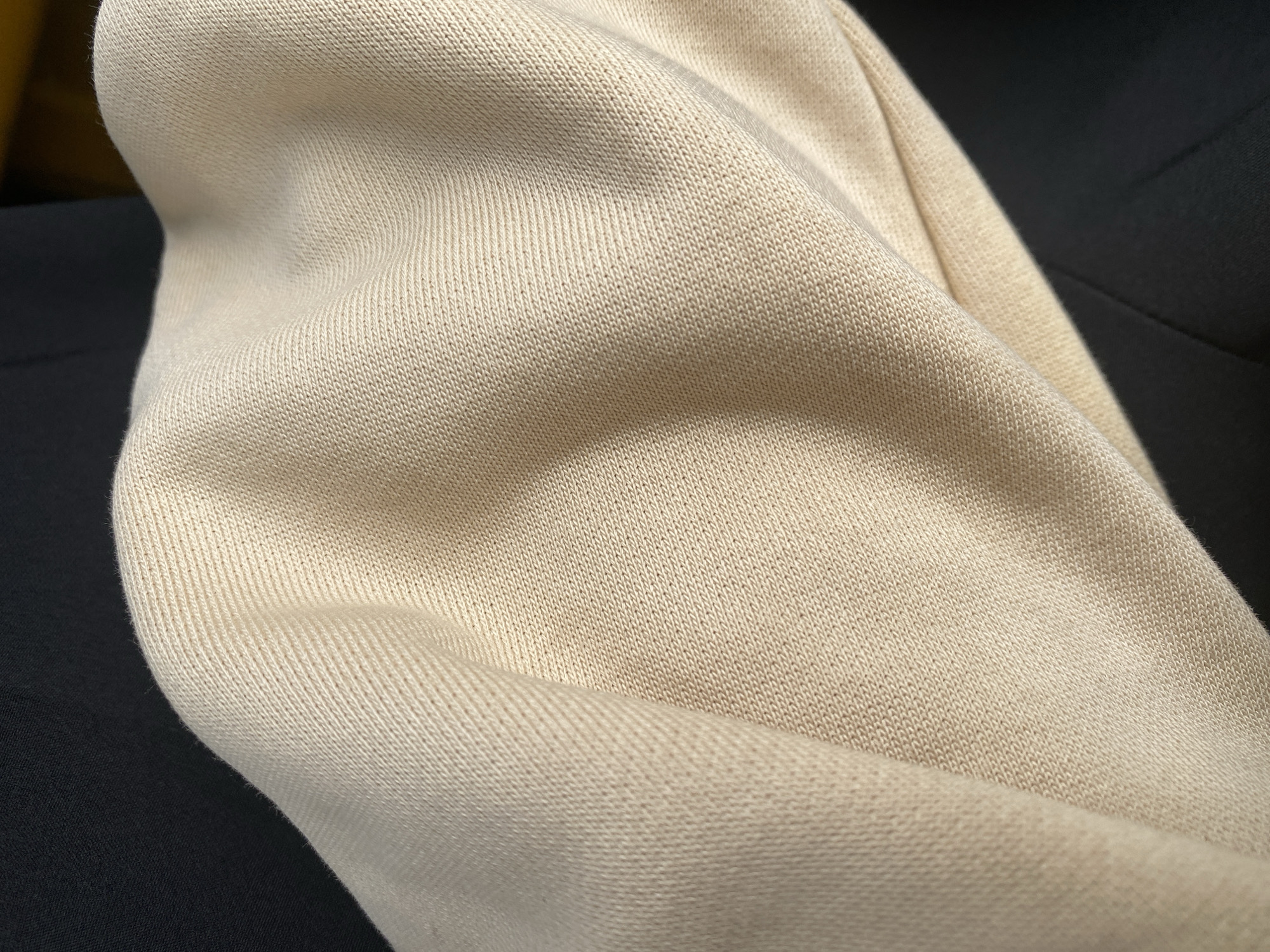 The media file shows a close-up, abstract view of draped beige fabric, likely clothing or upholstery, against a dark background. The fabric has a visible knit texture with small, repeating loops creating a ribbed appearance. Light falls across the fabric, highlighting the folds and creating subtle shadows that emphasize the material's softness and texture. The dark background is out of focus, suggesting it is a contrasting surface or perhaps a shadow. There are no people, identifiable objects, or discernible activities taking place. The scene is static and focuses entirely on the tactile and visual qualities of the fabric. The lighting suggests an indoor setting, possibly with diffused natural light or artificial lighting. No text is visible in the image. The context of Jalingo, Nigeria, does not provide any specific visual cues within the frame itself, as the image is an abstract close-up of fabric.