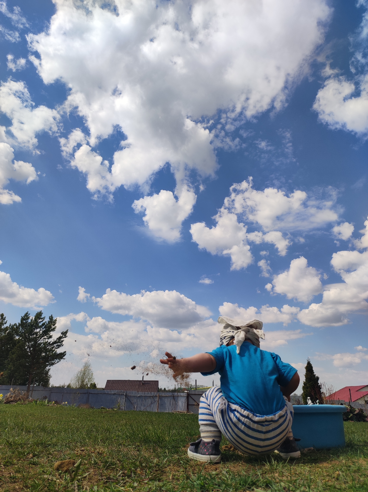 A child wearing a blue shirt, striped pants, and a white head covering squats in a grassy yard, throwing dirt into the air with their right hand. The scene is outdoors under a bright blue sky dotted with fluffy white clouds. A tree is visible to the left, and houses with red roofs and a fence are in the background. A blue container sits behind the child. The location is Фоки, Russia.