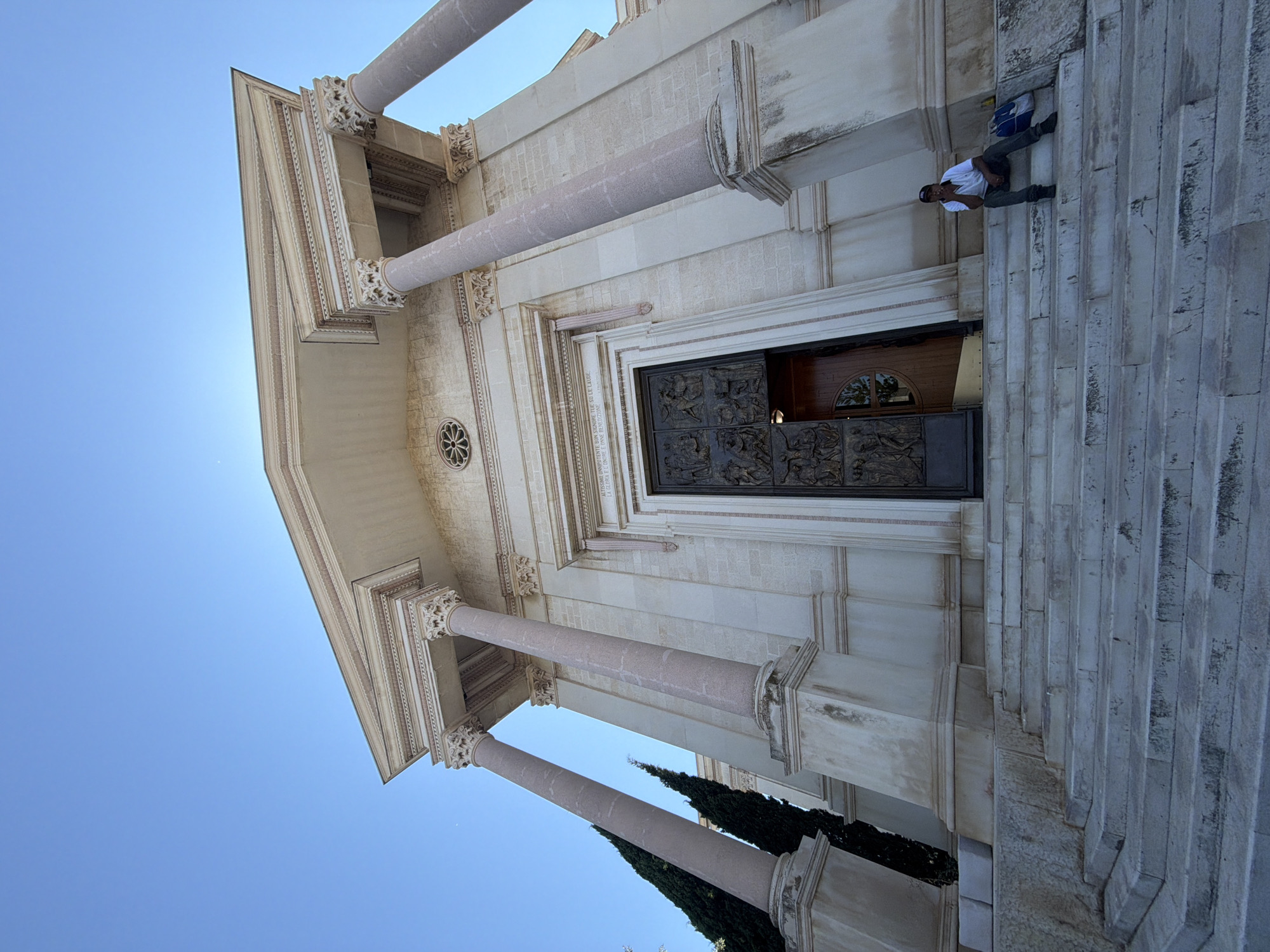 A wide-angle, low-angle shot depicts the Basilica di Santa Croce in Bari, Italy, under a clear blue sky. The facade of the basilica features grand classical architecture, with large pinkish-grey columns supporting a pediment. The entrance is an ornate, dark metal double door, partially ajar, revealing a glimpse of the interior. To the right of the entrance, a man wearing a white polo shirt, dark pants, and a baseball cap is seated on the broad, stone steps leading up to the basilica. He is holding a cigarette and appears to be looking to his left. Next to him on the steps is a blue backpack. Two tall, dark green cypress trees stand to the left of the basilica's entrance. The steps and the building's lower facade show signs of age and wear, with some staining and discoloration. The overall scene is one of architectural grandeur contrasted with a moment of quiet repose.