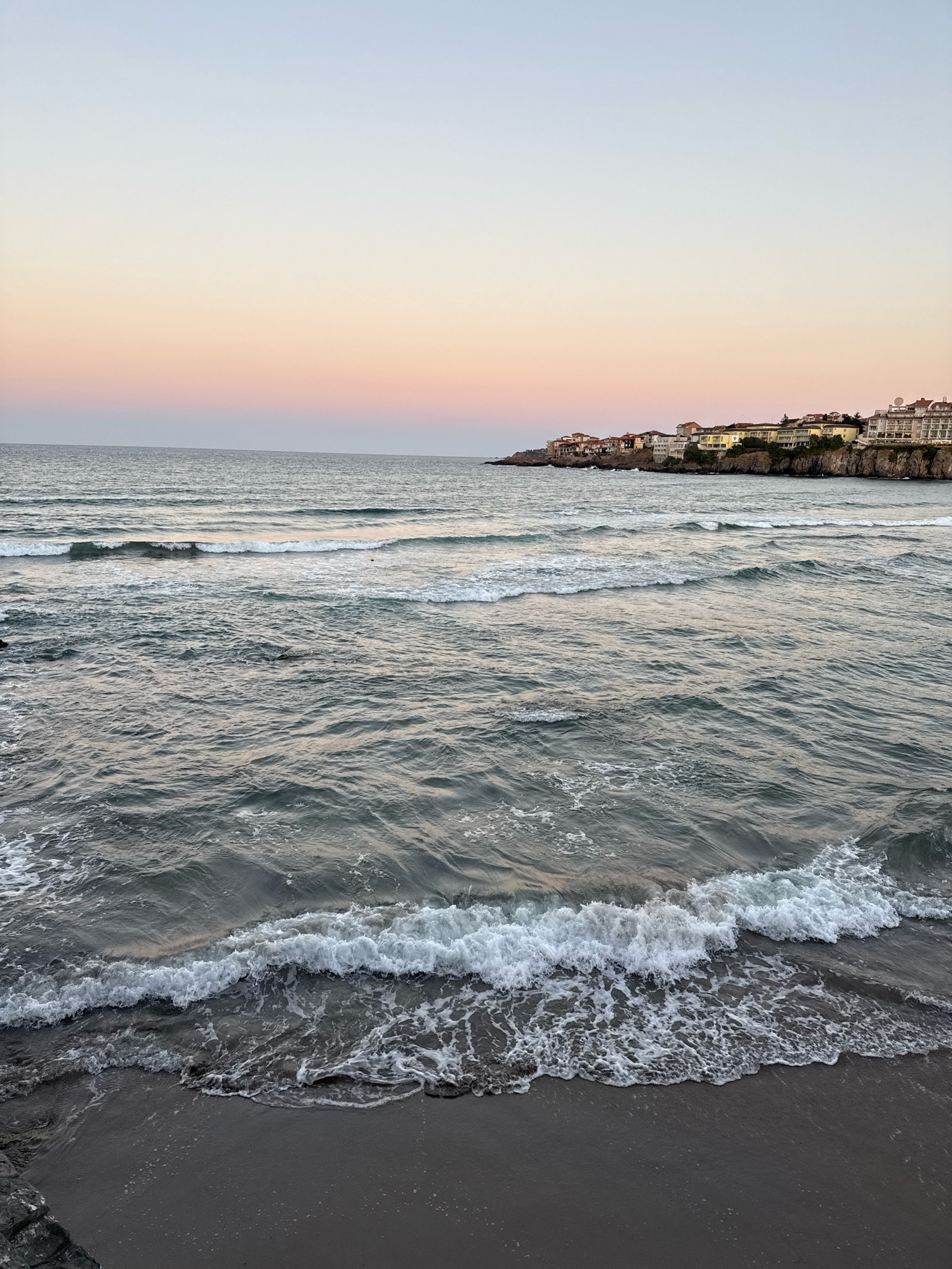 A wide-angle, eye-level shot captures the sunset over the Black Sea coast in Sozopol, Bulgaria. The sky transitions from a pale blue at the top to soft hues of pink and orange closer to the horizon. The sea is calm with gentle waves rolling towards the shore, creating white foam as they break on the dark sand. In the background, a cliff face is lined with buildings, characteristic of Sozopol's old town, suggesting a coastal town setting. There are no people or animals visible in the frame. The overall mood is serene and tranquil, with natural lighting from the setting sun.