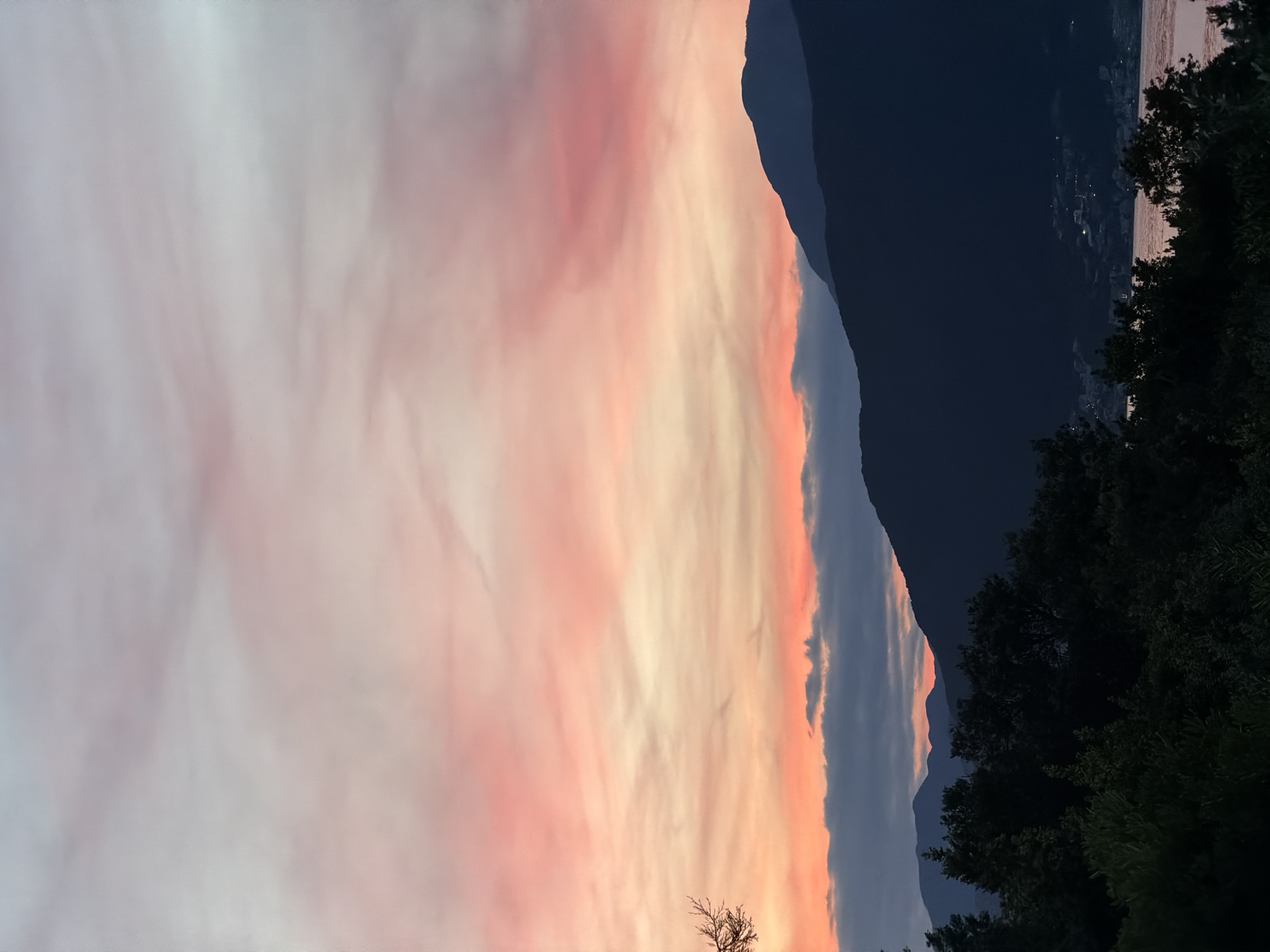 The image shows a landscape in Gošići, Montenegro at dusk. The sky transitions from a pale, almost white hue on the left, to vibrant shades of pink and orange. A dark mountain range stretches across the middle, adjacent to the sunset colors. To the right of the mountain, a body of water reflects faint city lights. Dark green foliage, possibly trees and bushes, occupies the right foreground.