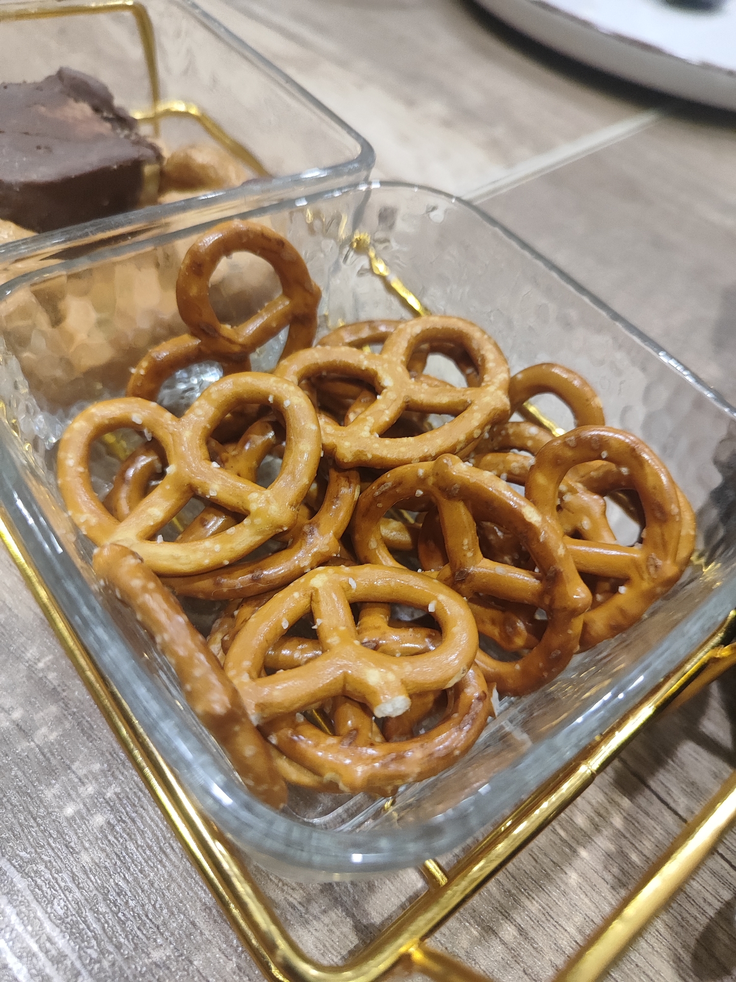 The image shows a square glass dish filled with mini pretzels. The pretzels are golden brown and sprinkled with salt. The dish is supported by a gold-colored metal stand. In the background, slightly out of focus, is another glass dish containing a dark, rectangular confection and some lighter colored items, possibly nuts or other candies. The scene is set on a textured surface that appears to be a table. The image is taken from a slightly elevated angle, looking down into the dish of pretzels. The overall impression is that of a snack or appetizer presentation.