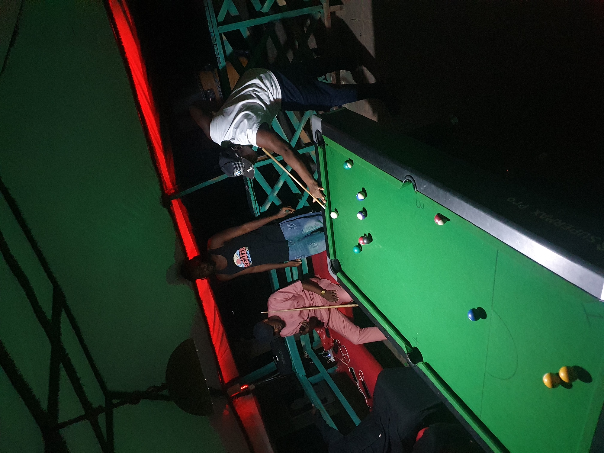 Three individuals are playing pool on a green pool table under low lighting conditions. The location appears to be outdoors. The people are gathered around the pool table. One person is actively playing the game while the other two appear to be spectating. The environment seems dimly lit.