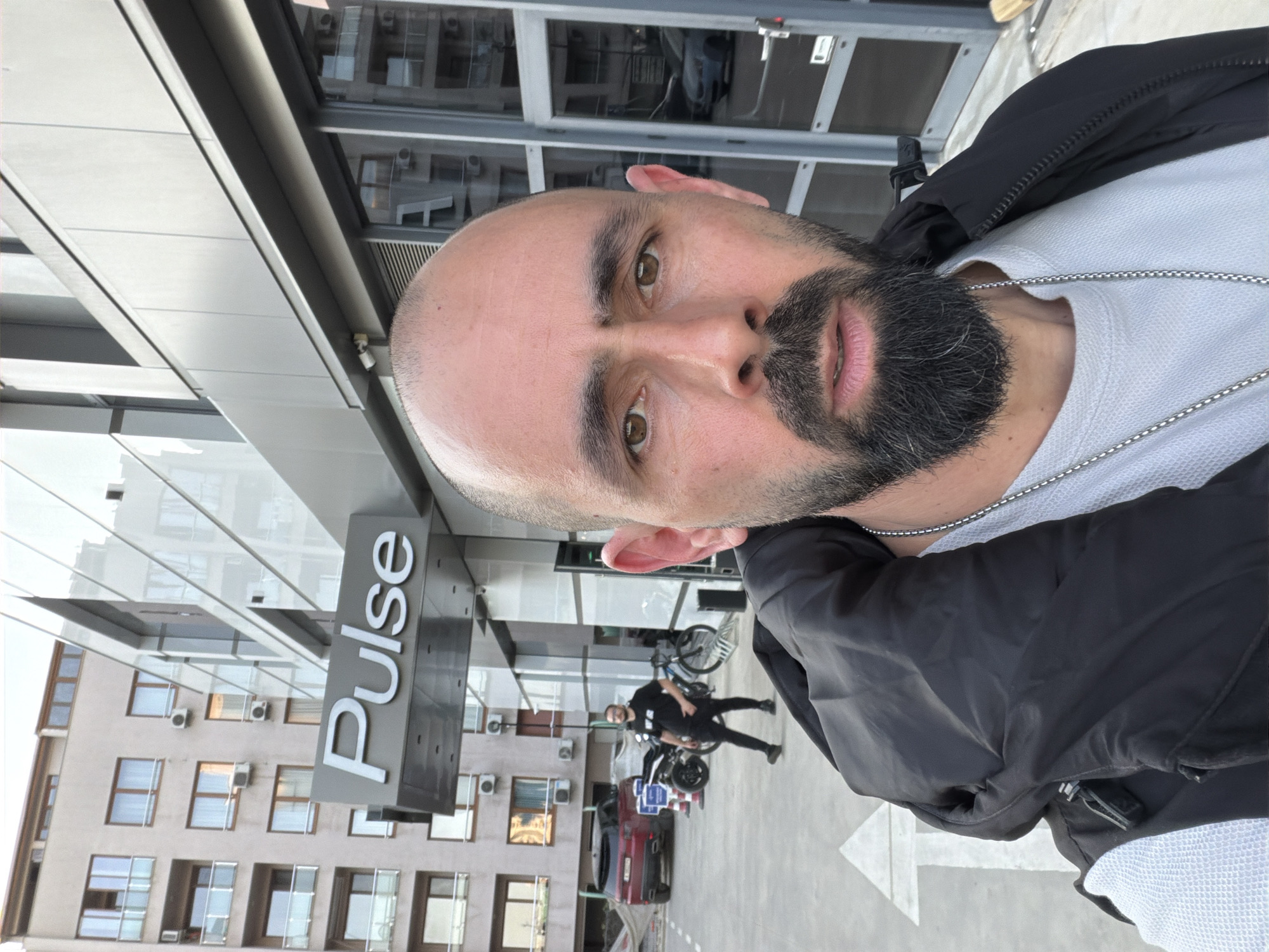 The image depicts a man with a shaved head and a beard, wearing a light-colored shirt and a black jacket. He is standing outside a building with the sign "Pulse" on it. In the background, there is a multi-story building with numerous windows and air conditioning units. A man on a motorcycle can be seen in the background. This scene is likely in Plovdiv, Bulgaria, given the presence of the "Pulse" sign which is a known location in the city.