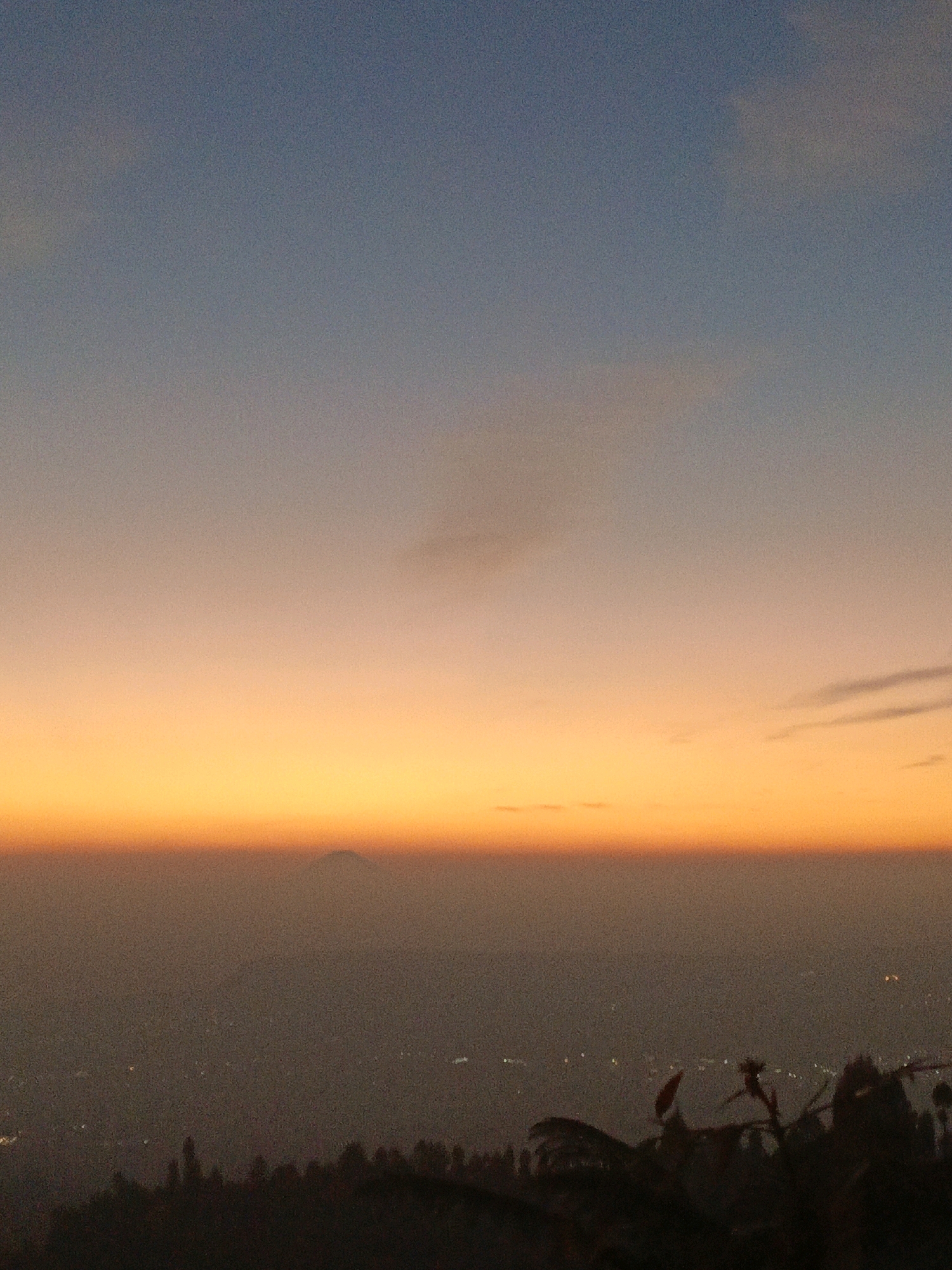 This image depicts a sunset over a hazy landscape, with a distinct mountain peak visible in the distance. The sky transitions from a deep blue at the top to warm hues of orange and yellow near the horizon. The foreground shows a dark silhouette of trees and vegetation. Lights from what appears to be a town or city are faintly visible in the middle ground, partially obscured by the haze. The location is identified as Butuh Kidul, Indonesia. The scene is tranquil and atmospheric, capturing the subdued light of dusk.